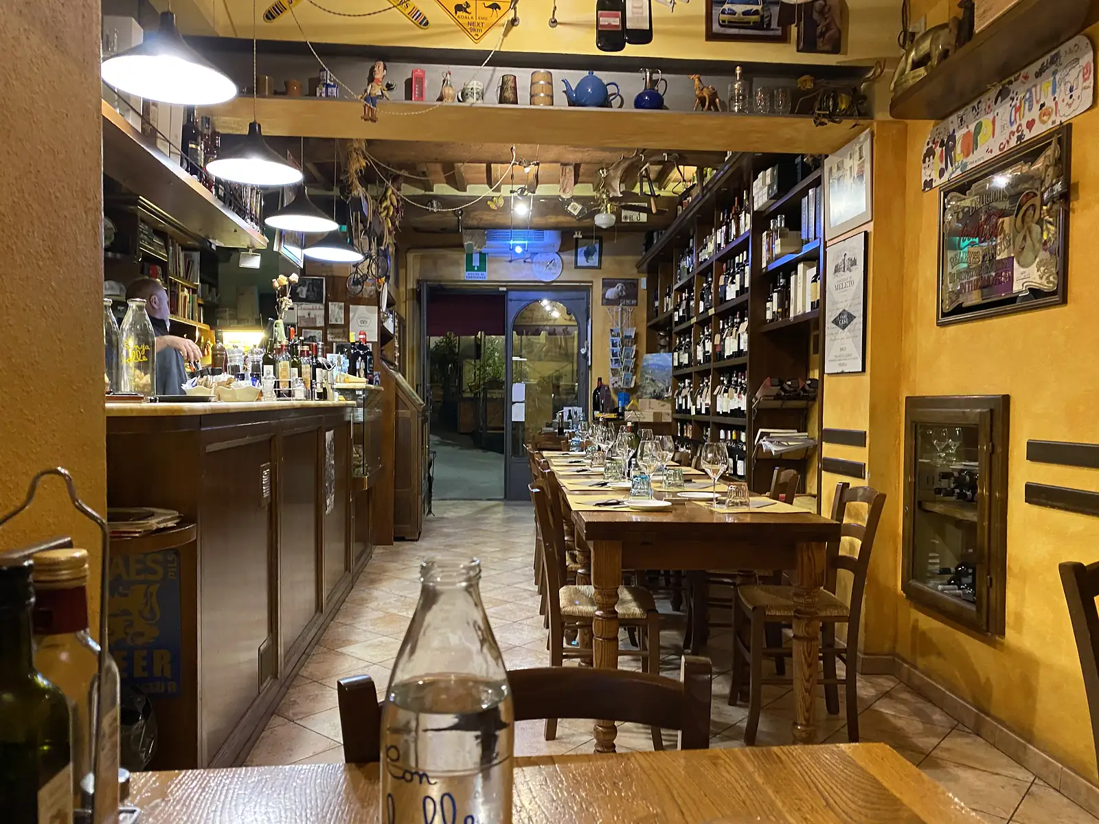 The interior of a rustic Italian trattoria with laid tables and wine glasses beneath shelves crammed with wine bottles, and a barman behind the counter.