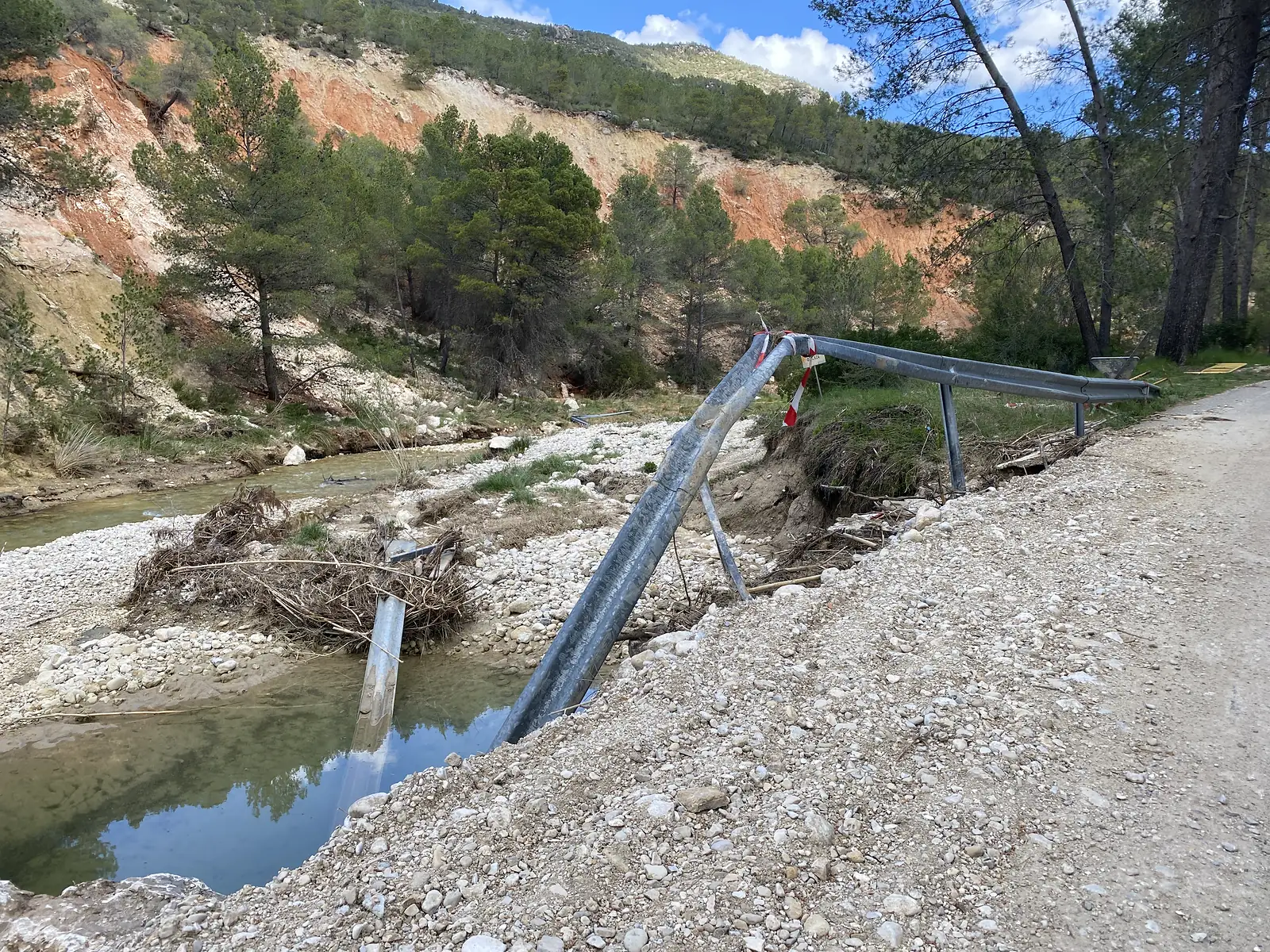 A crash barrier torn from its posts and bent down into a stony riverbed, red-and-white tape flapping from it and the road's edge scoured away.
