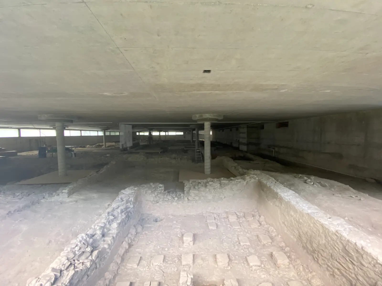 Excavated stone wall foundations of an archaeological dig lie exposed on a dusty floor beneath the concrete columns and ceiling of a modern building.