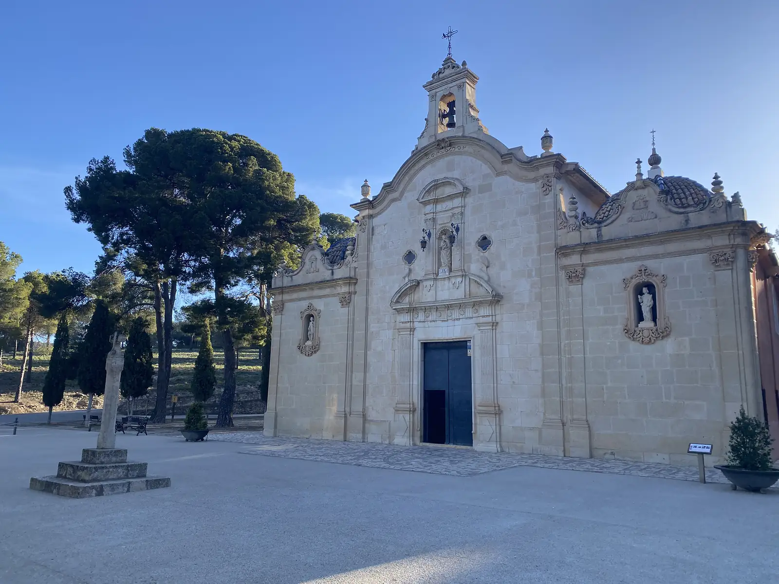 A baroque chapel with a scrolled gable, bell cote and statues in niches, a stone cross standing on the gravel forecourt.