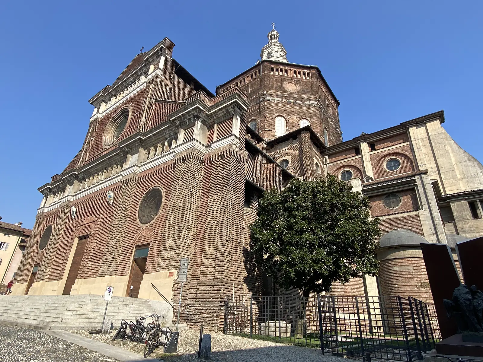 The vast brick façade and octagonal drum of Pavia's cathedral rise against a clear sky above its stepped entrance and a magnolia tree.