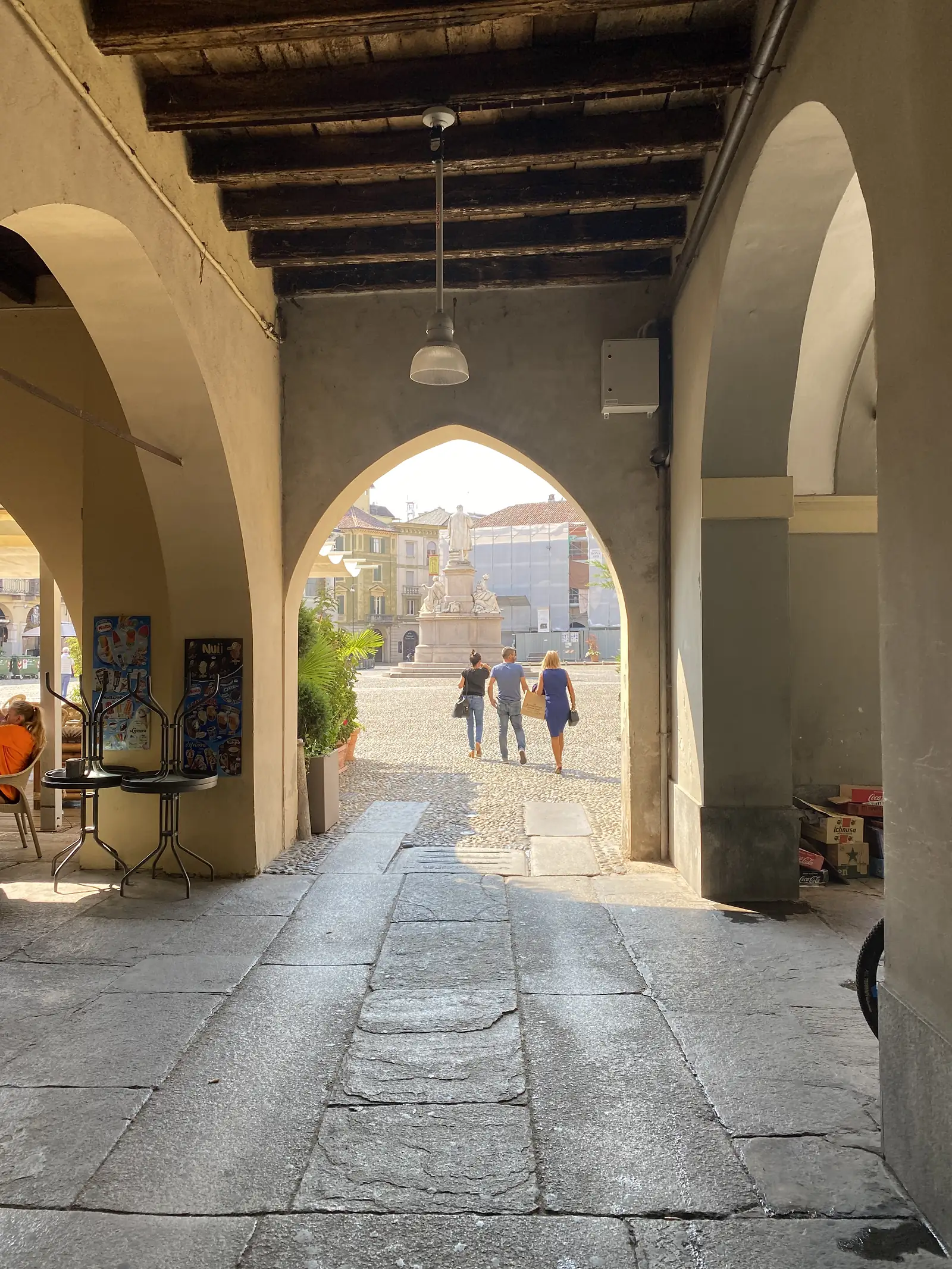Looking out through a stone archway onto a sunlit piazza where pedestrians walk past a fountain.