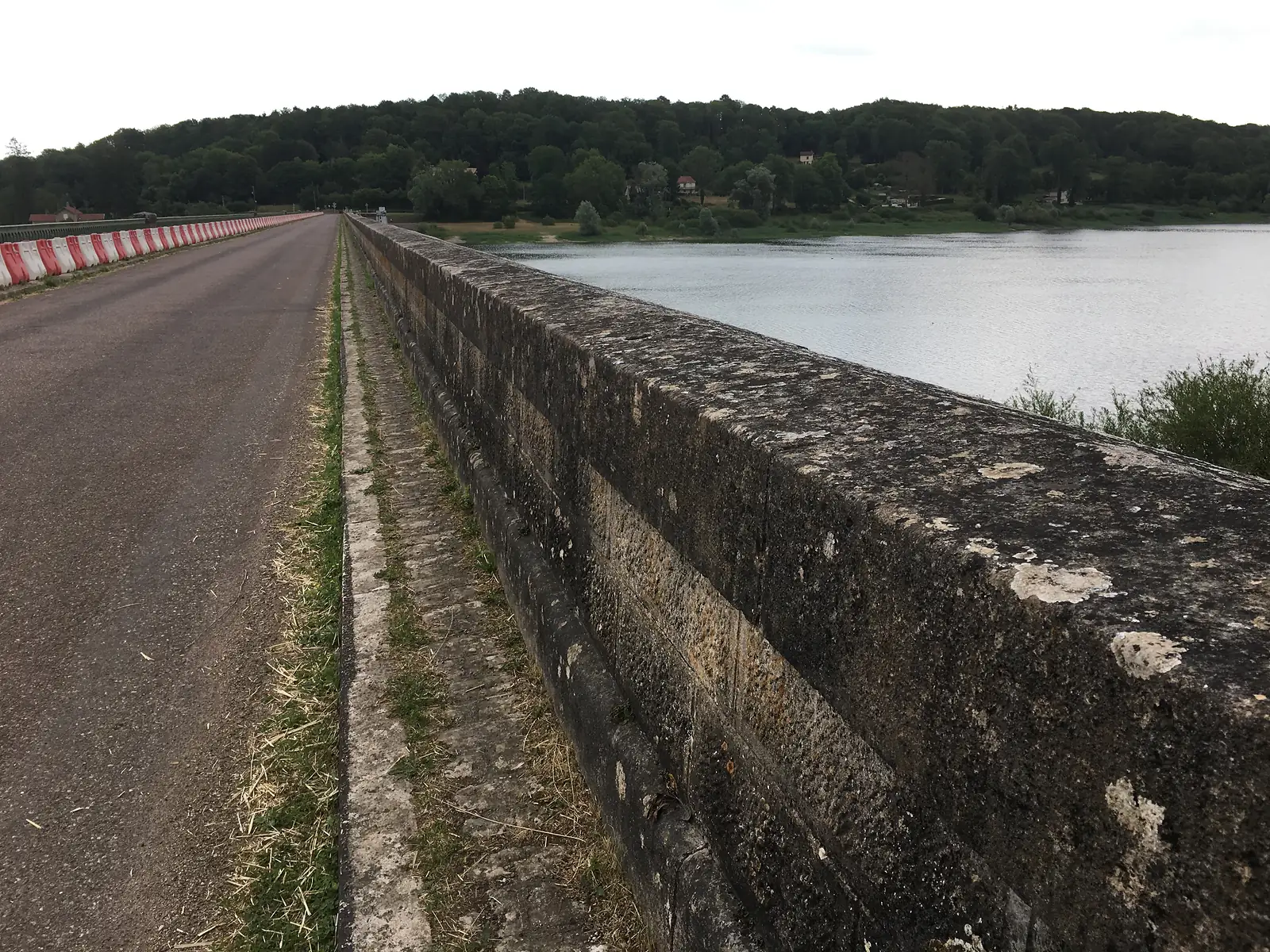 A stone causeway wall running alongside a reservoir, with a red-and-white barriered road on one side and wooded hills across the water.