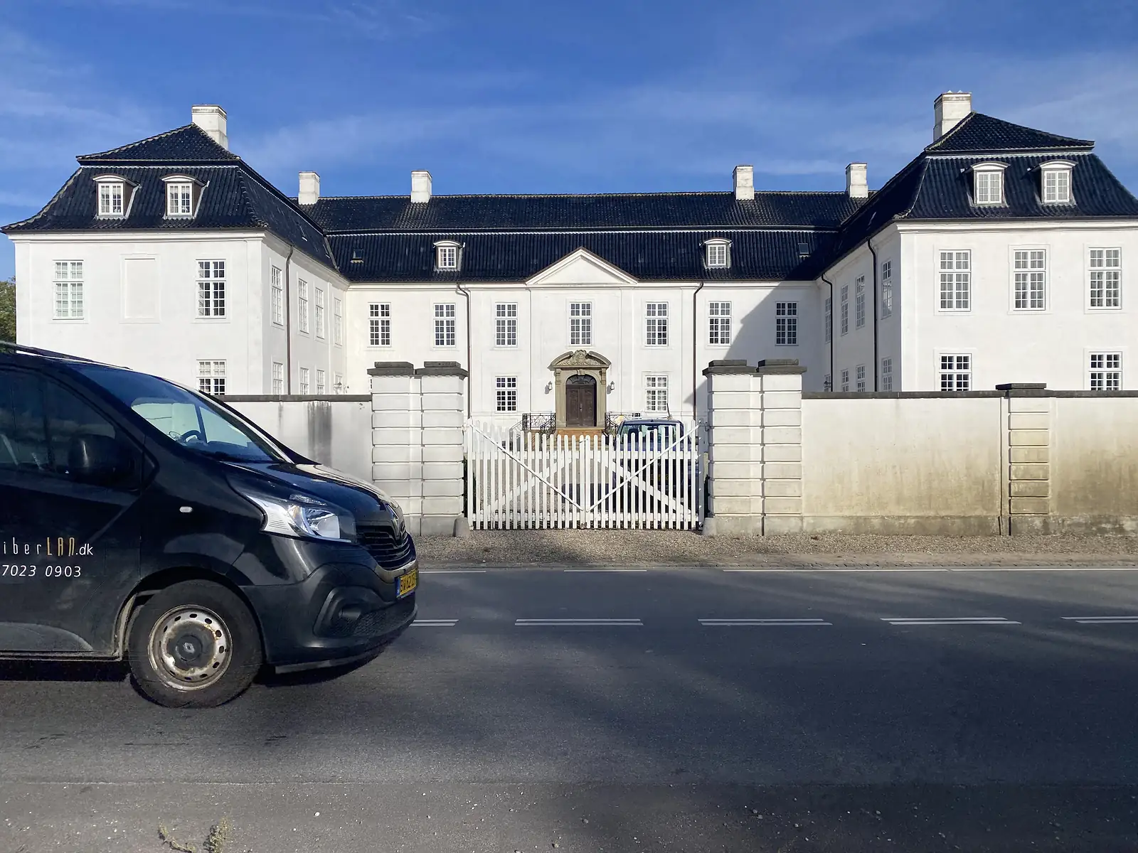 A long white manor house with a black mansard roof and dormers behind a white picket gate, a van passing on the road outside.