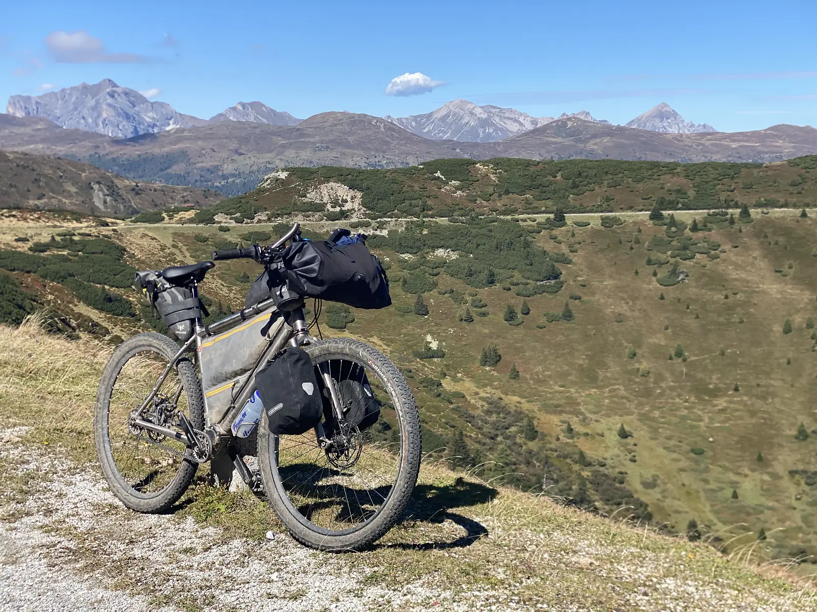 A loaded bikepacking bike stands on a stony shoulder above a bowl of dwarf pine, with a wide horizon of pale rocky peaks behind.