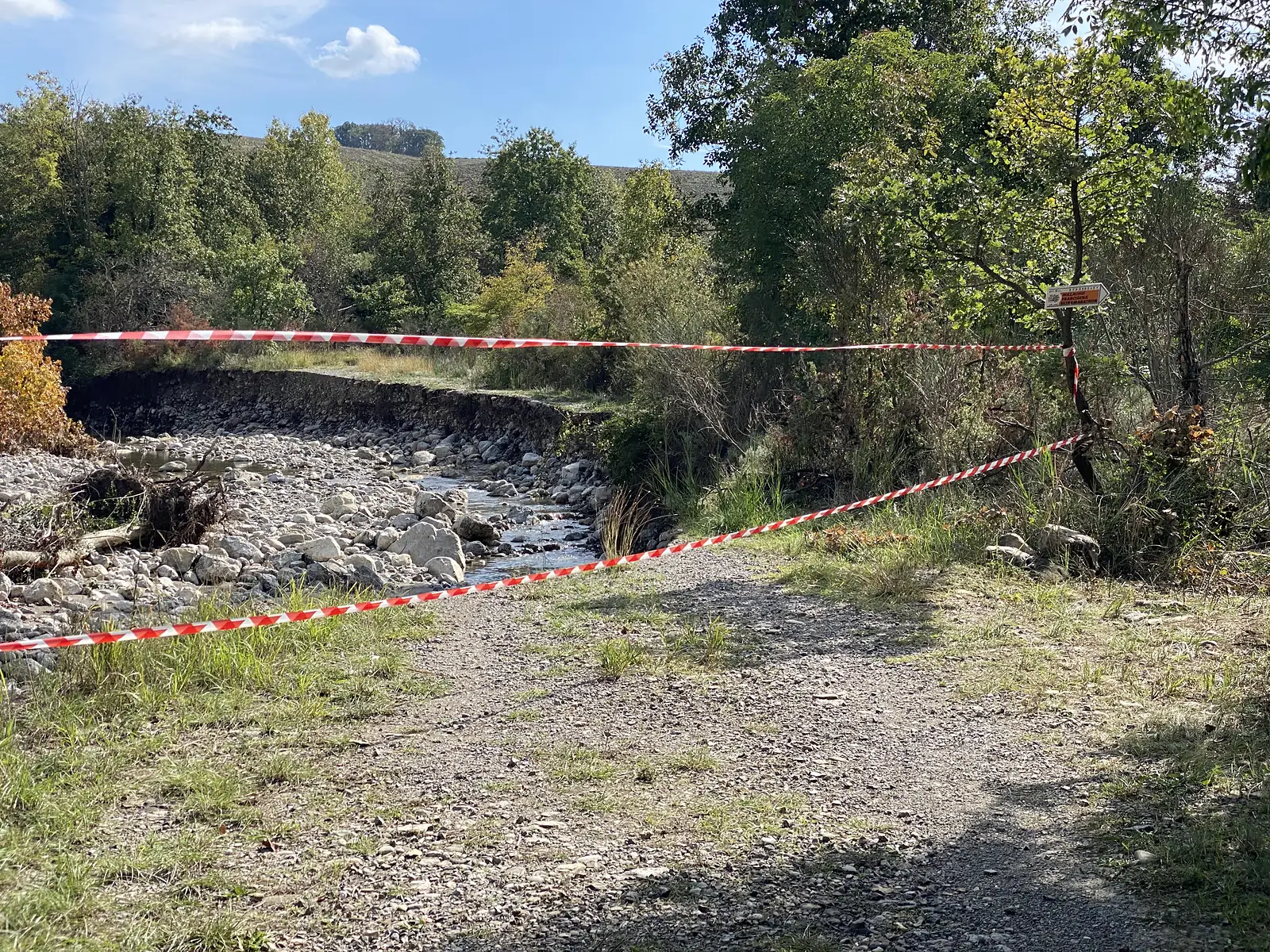 Red-and-white barrier tape strung across a gravel track next to a stony, mostly-dry riverbed, with a route signpost among trees on the right.