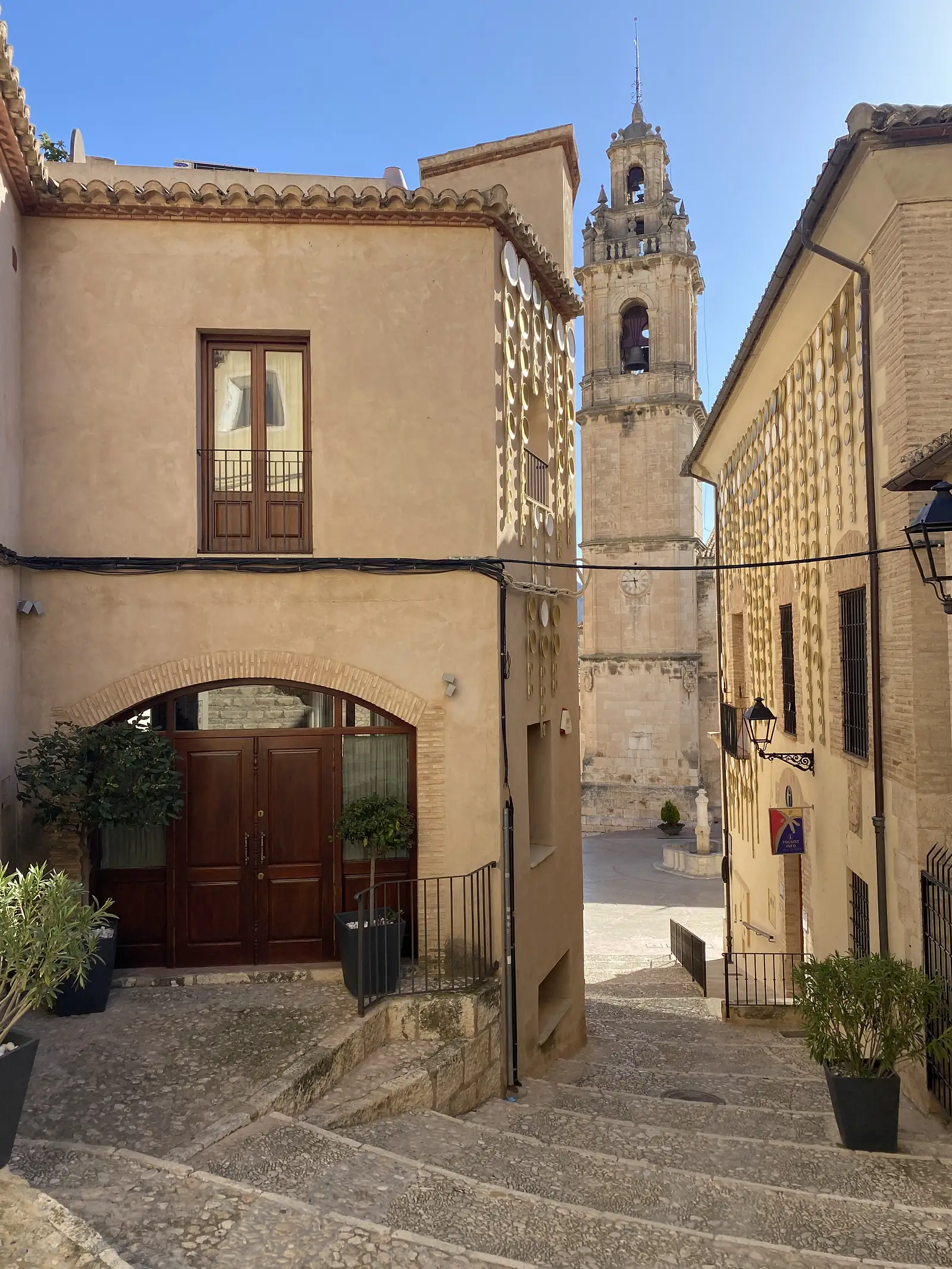 A cobbled stepped street between ochre houses leading towards an ornate stone bell tower with a clock.