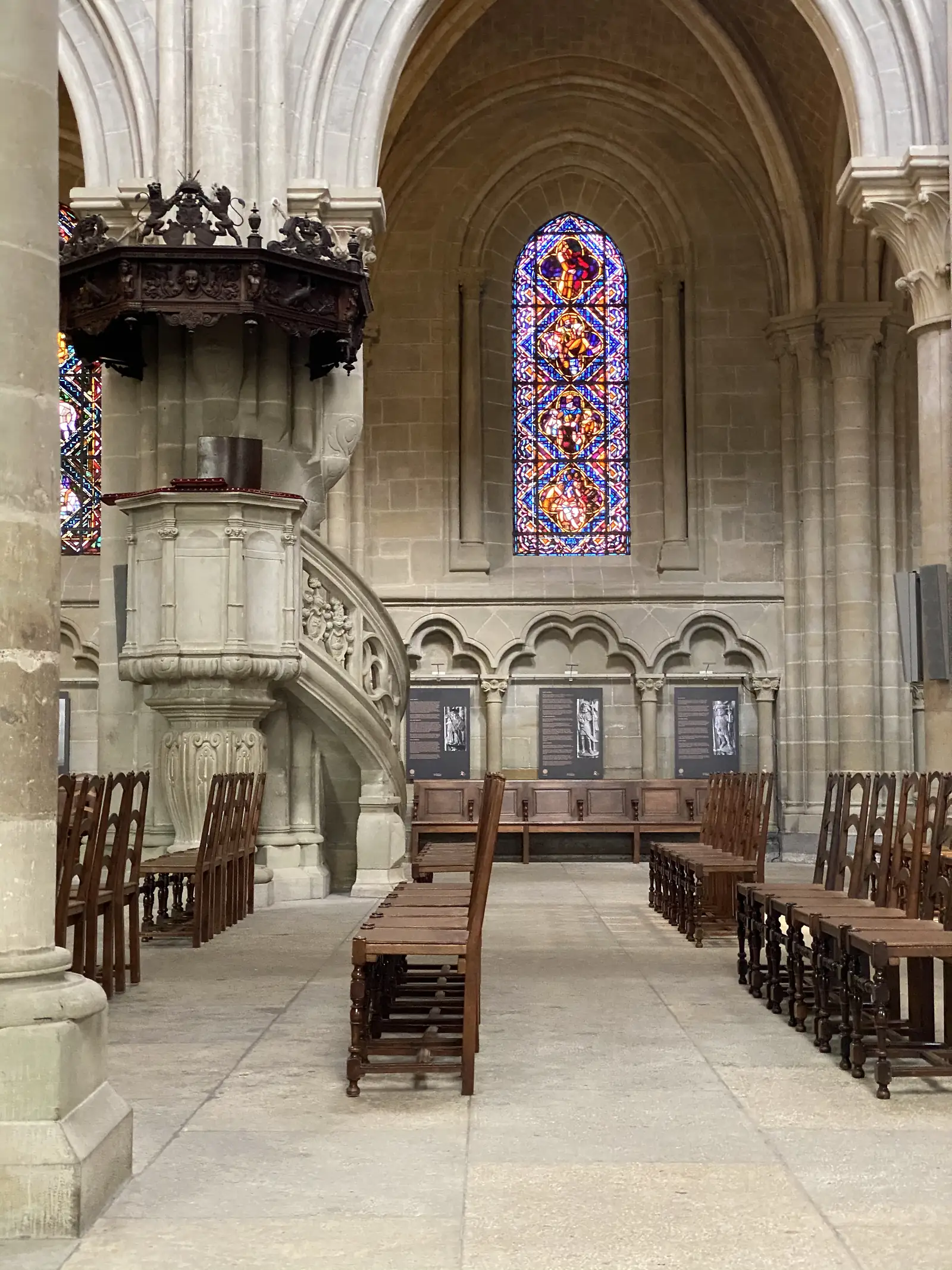 An ornate carved stone pulpit with a spiral stair beneath a tall stained-glass window inside Lausanne Cathedral, rows of wooden chairs facing the aisle.