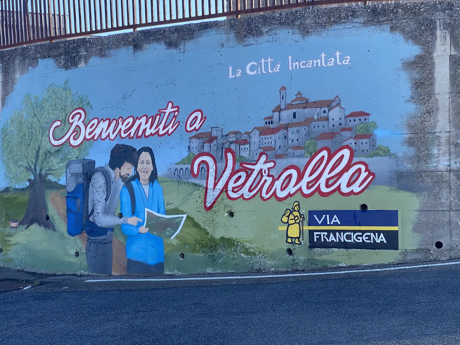 A painted mural under a road bridge showing two hikers reading a map beside a hilltop town, with the words "Benvenuti a Vetralla", "La Città Incantata" and a Via Francigena pilgrim waymark logo.