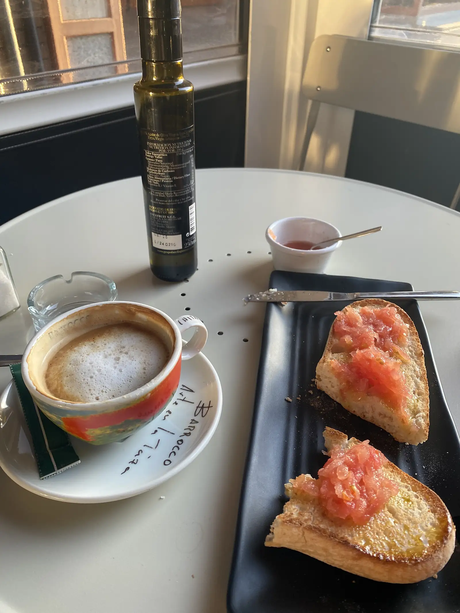 Two slices of toast spread with grated tomato on a black plate, beside a milky coffee and a bottle of olive oil on a cafe table.