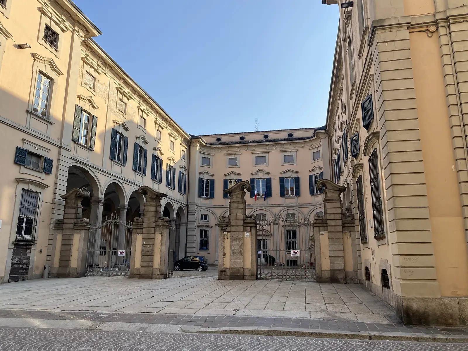 Wrought-iron gates open into a grand courtyard flanked by an arcaded loggia and shuttered palazzo façades.