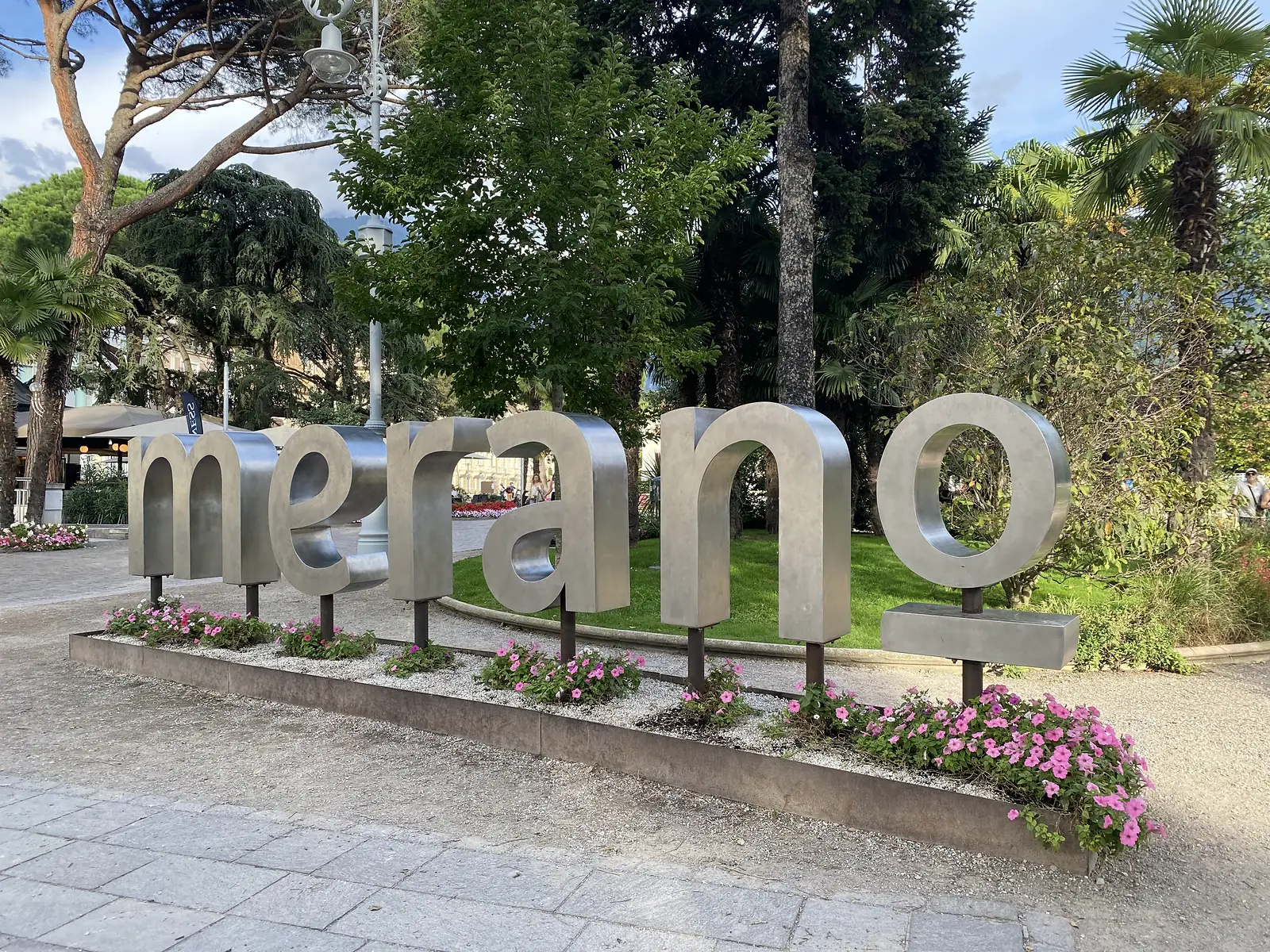 Large brushed-metal letters spelling "merano" stand in a bed of pink petunias in a park of pines and palms.