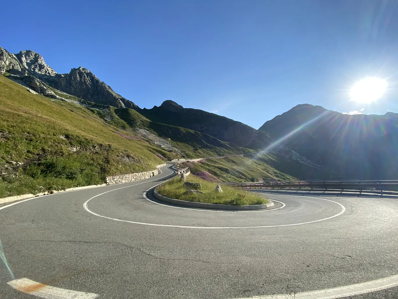 A sharp hairpin bend on a mountain road, with the morning sun flaring low over a ridge and pink alpine flowers growing along the tarmac's edge.