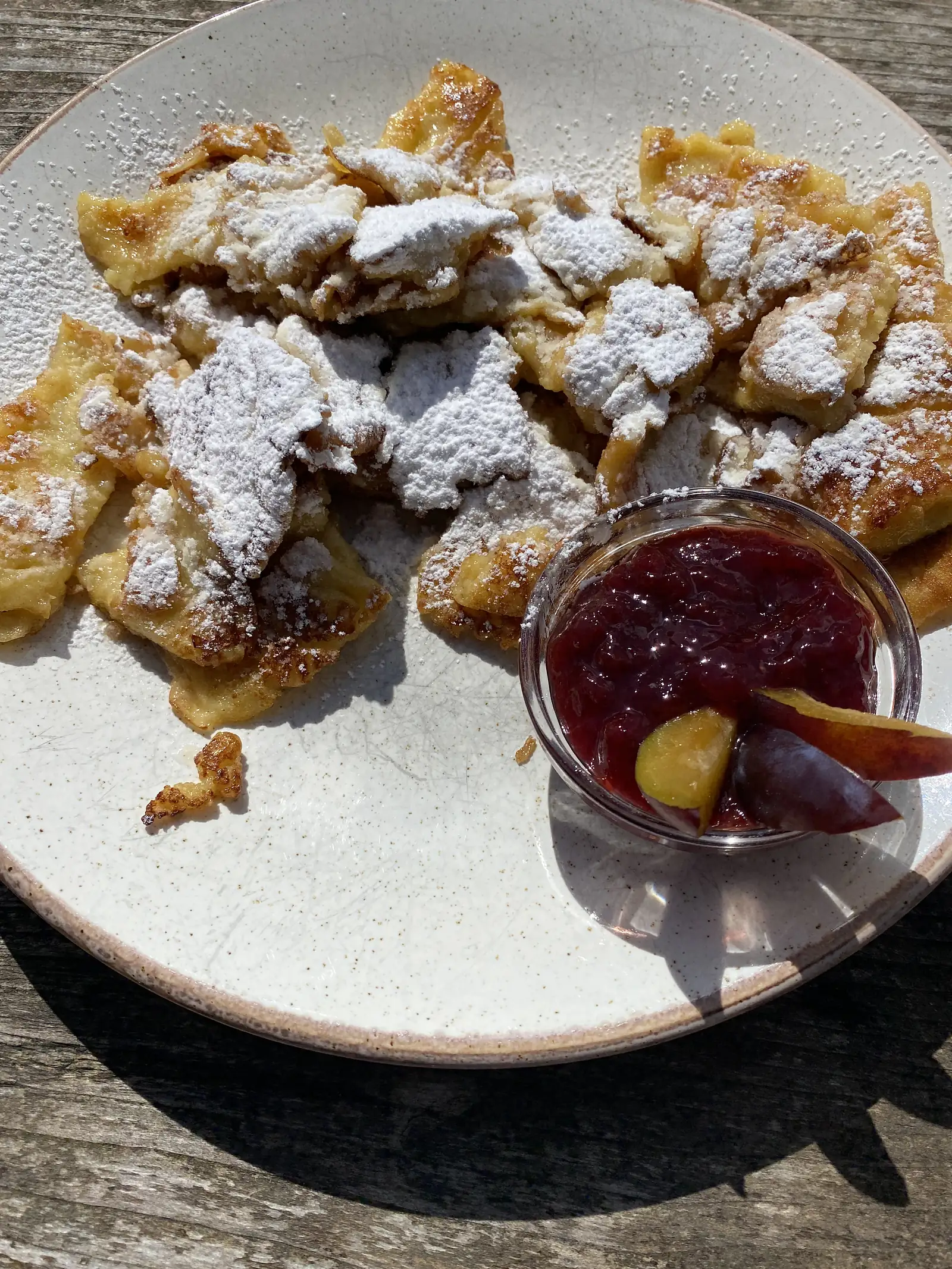 A plate of torn, sugar-dusted pancake pieces with a glass dish of dark plum compote, on a weathered wooden table in the sun.