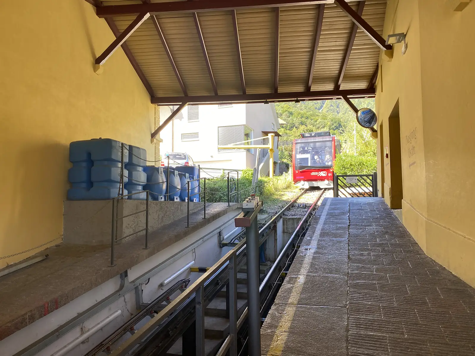A red funicular car waits at the top of its incline beneath a yellow-walled station canopy, cable and rails running away downhill.