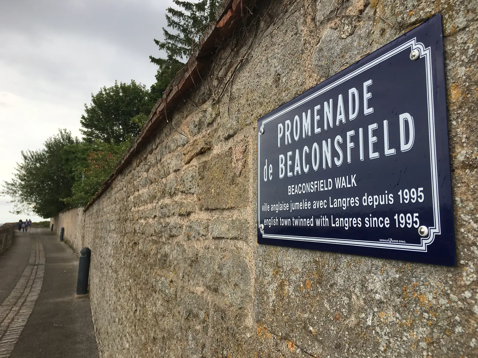 A stone plaque on the Langres ramparts reading Promenade de Beaconsfield, Beaconsfield Walk, noting the English town twinned with Langres since 1995, with walkers on the path beyond.
