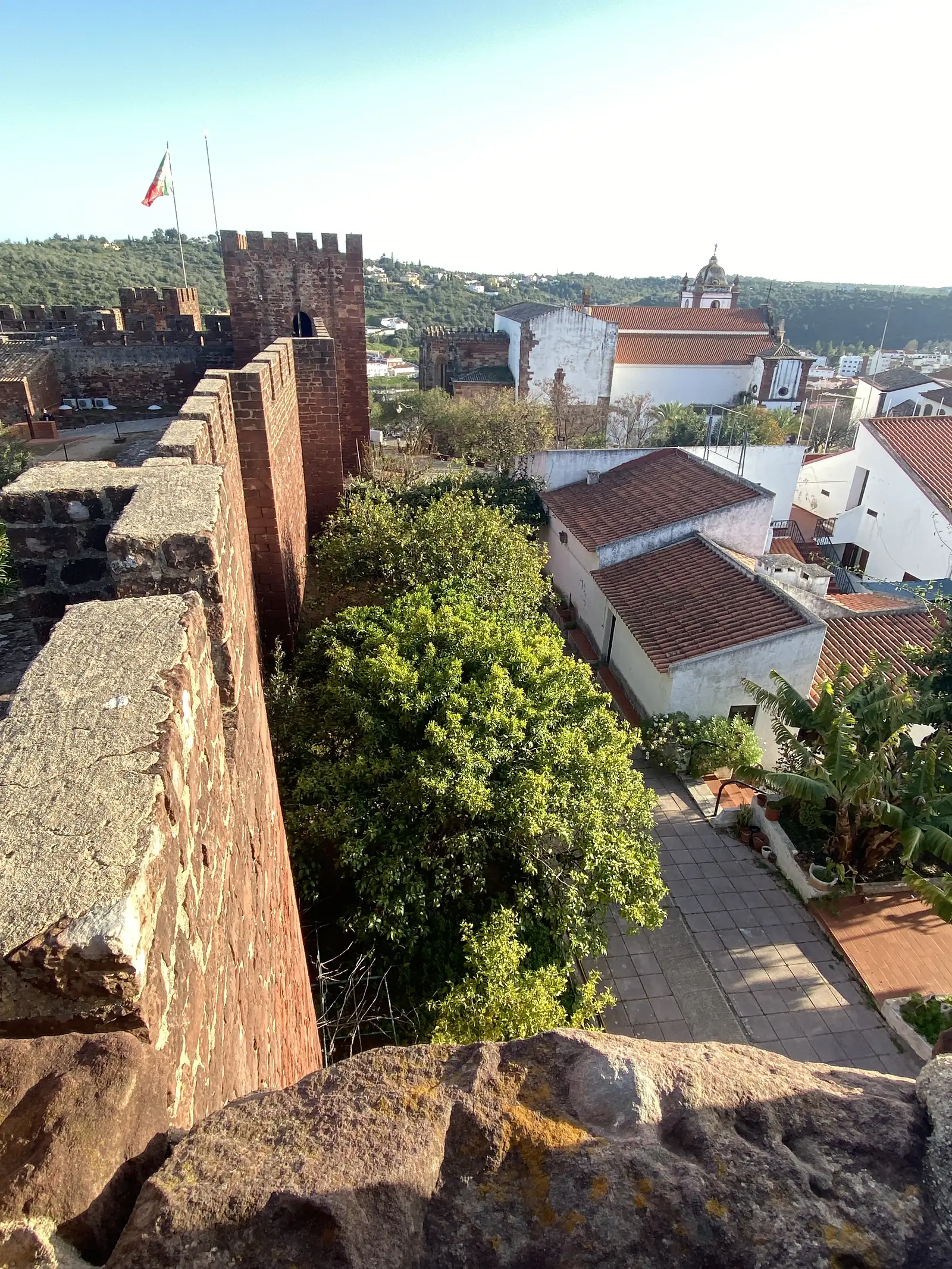 Looking down from a castle rampart along the outside of its red sandstone wall to the terracotta roofs and whitewashed houses of the town below, with the Portuguese flag flying from a crenellated tower and a church dome on the hillside beyond.