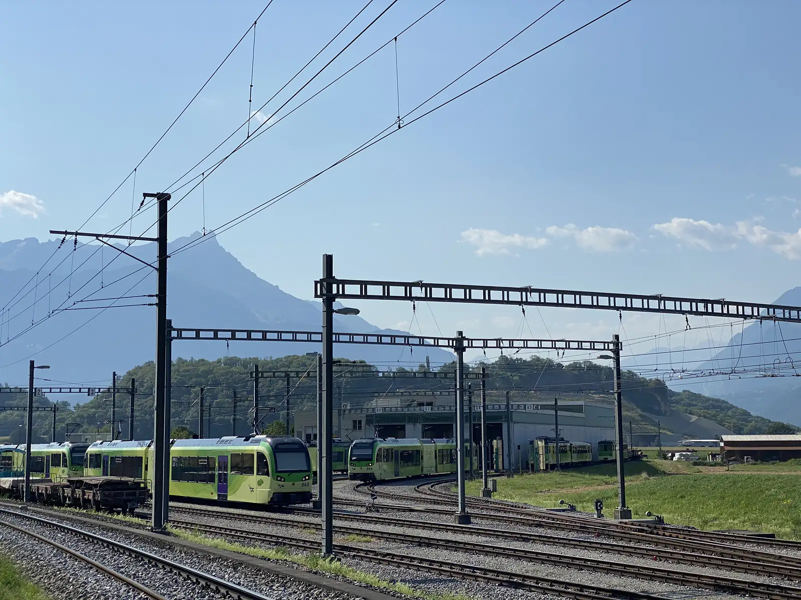 Green-and-white light-rail trains stand on tracks beneath overhead power gantries at a depot, mountains rising behind.