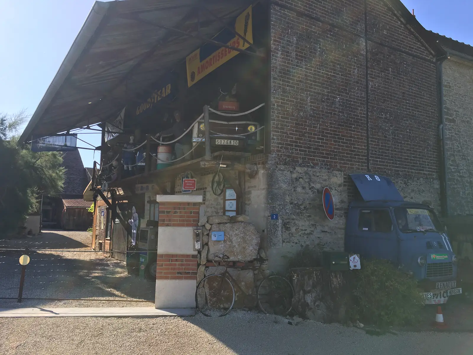 A ramshackle roadside garage crammed with vintage motoring memorabilia, including an old Renault van, mannequins, tyre adverts and a bicycle propped against the stone wall.