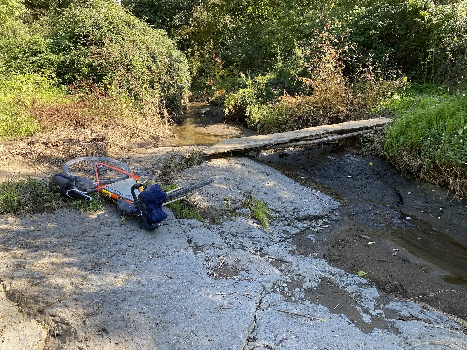A loaded bike lies on its side beside a shallow stream crossing, with a narrow wooden plank bridge spanning the water nearby.