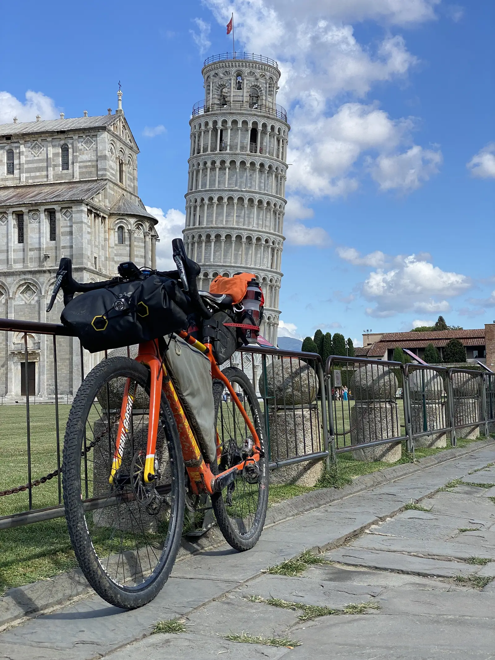 A laden gravel bike with orange bags and a bar bag leaning against a railing, the Leaning Tower of Pisa rising behind it against a blue sky.
