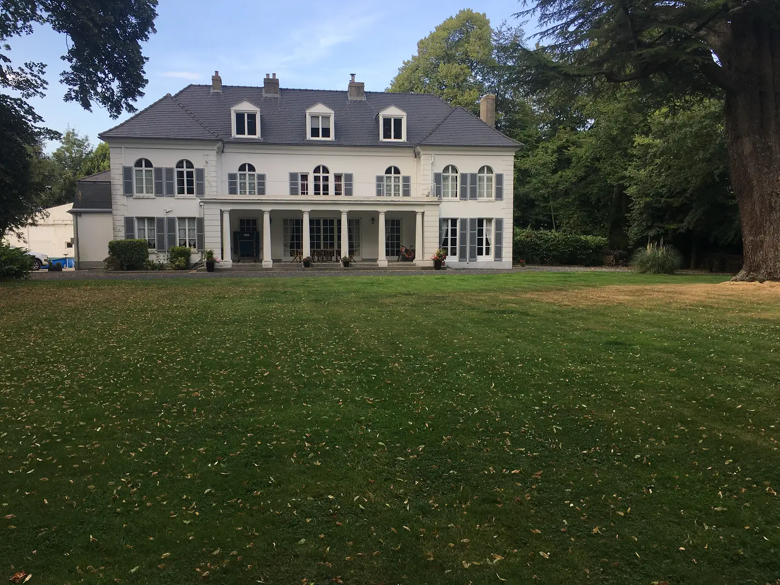 The rear facade of a grand manor house with a pillared terrace overlooking a wide lawn, a bike leaning against one of the columns.