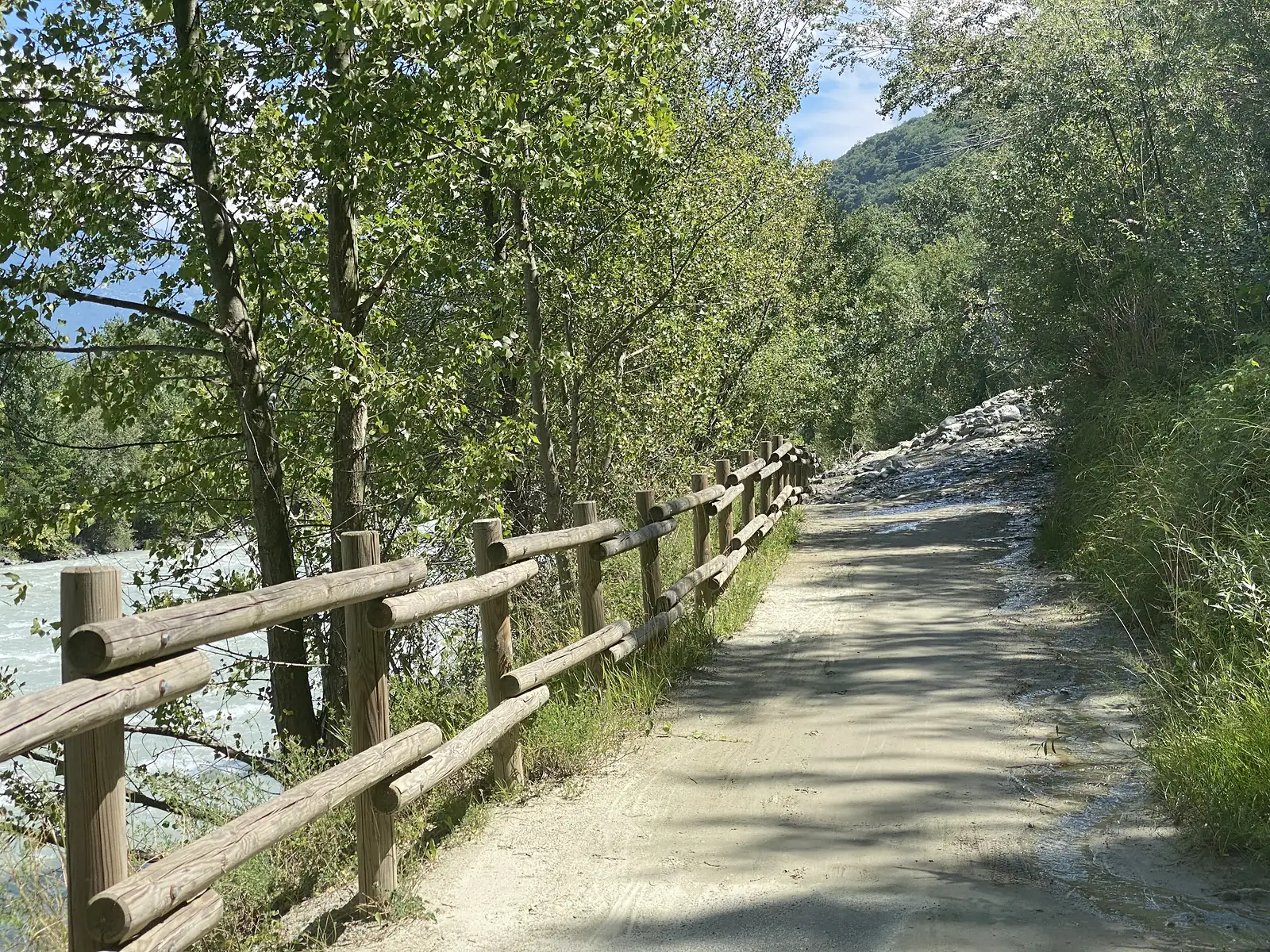 A gravel riverside path runs behind a post-and-rail wooden fence, poplar trees screening the pale turquoise glacial river flowing alongside.