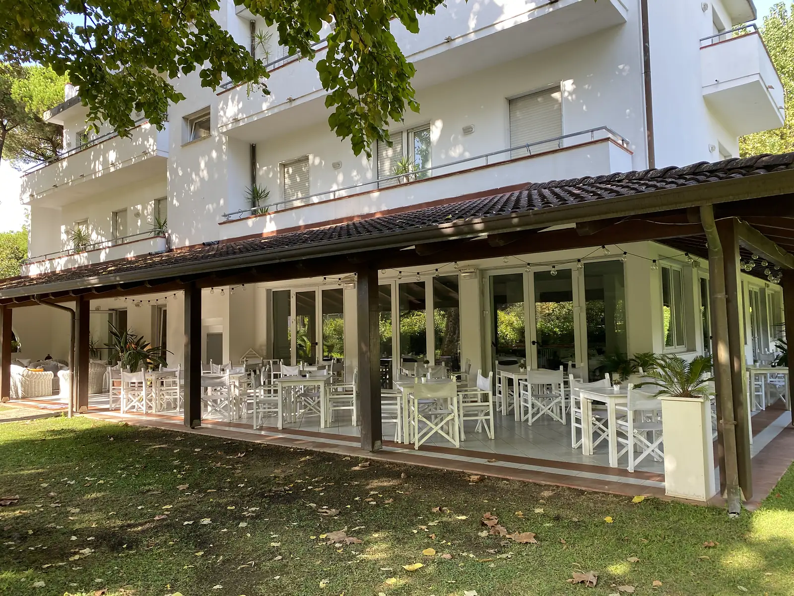 An open-sided dining terrace with white director's chairs and tables under a tiled pergola roof, set in front of a white apartment building shaded by trees.