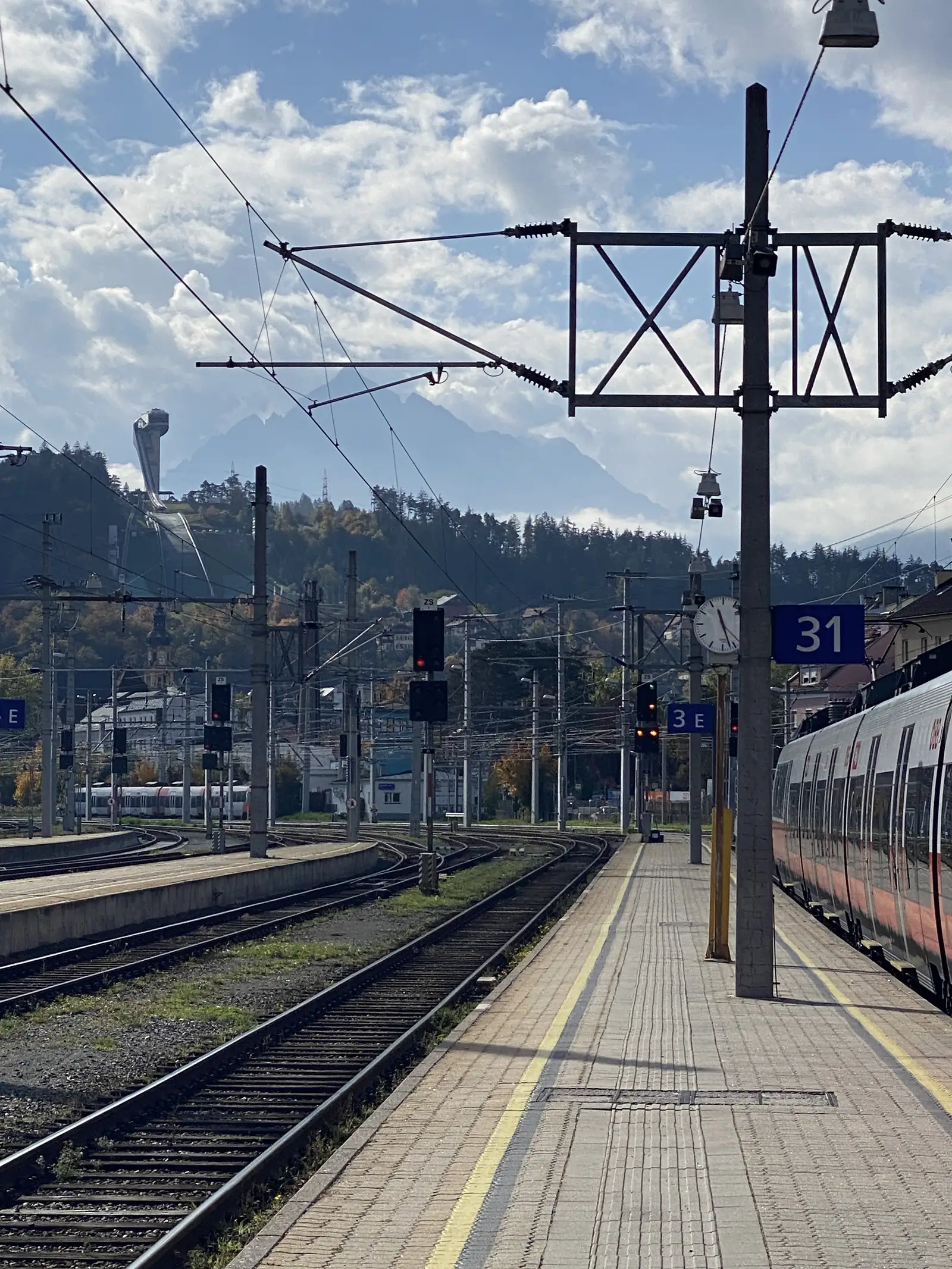 An open railway platform numbered 31 stretches beneath overhead signal gantries and power lines, with mountains visible in the distance.