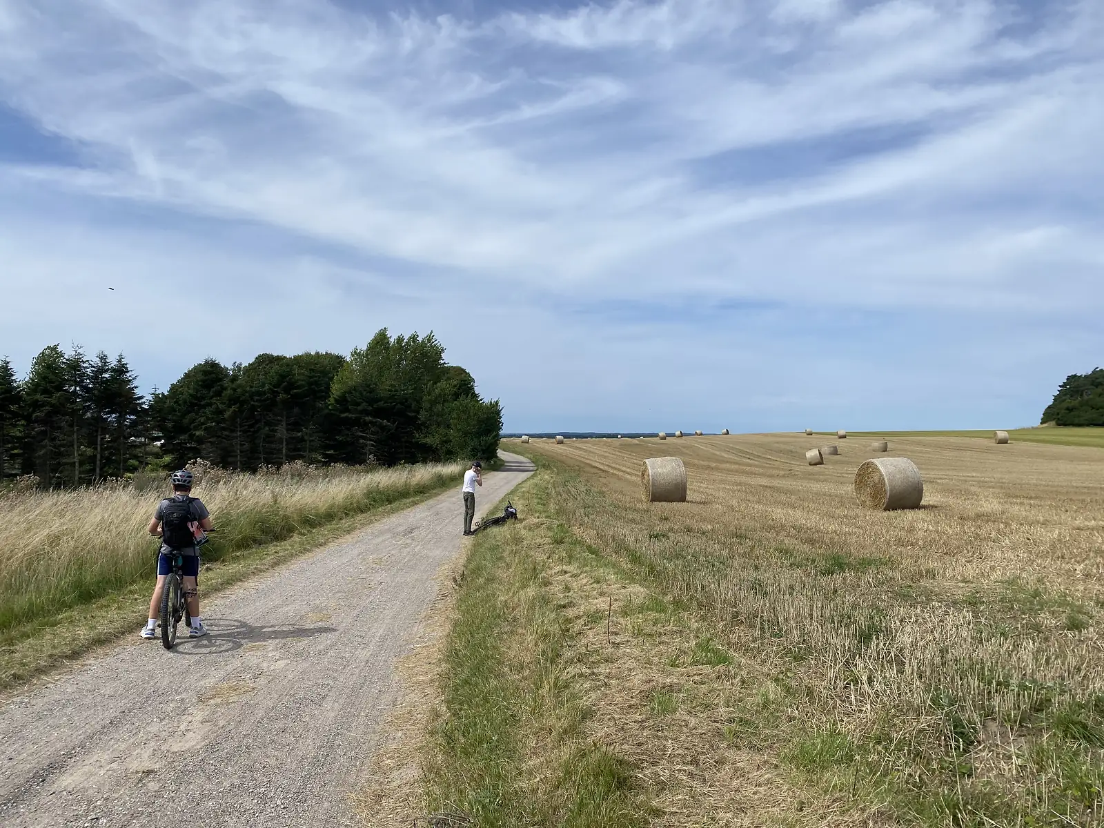 A cyclist on a gravel track beside a harvested field dotted with round straw bales, under a wide blue sky.