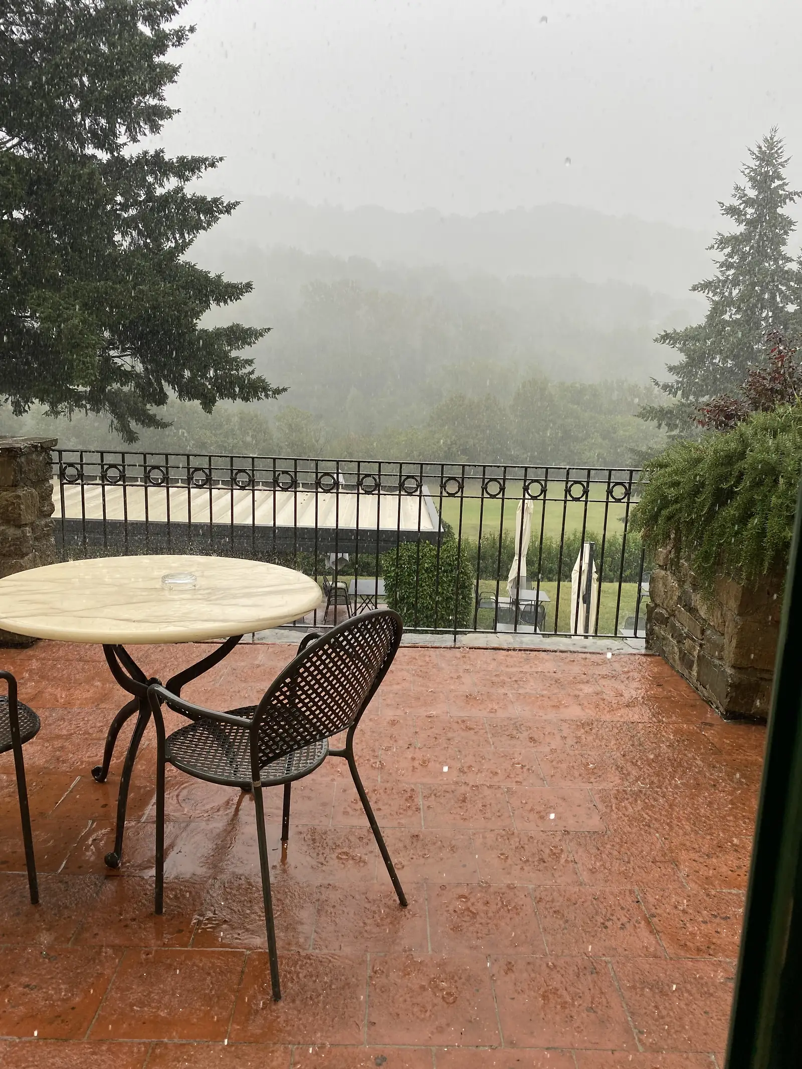An empty café table and chairs sit on a rain-speckled terrace behind a wrought-iron railing, mist obscuring the hills beyond.