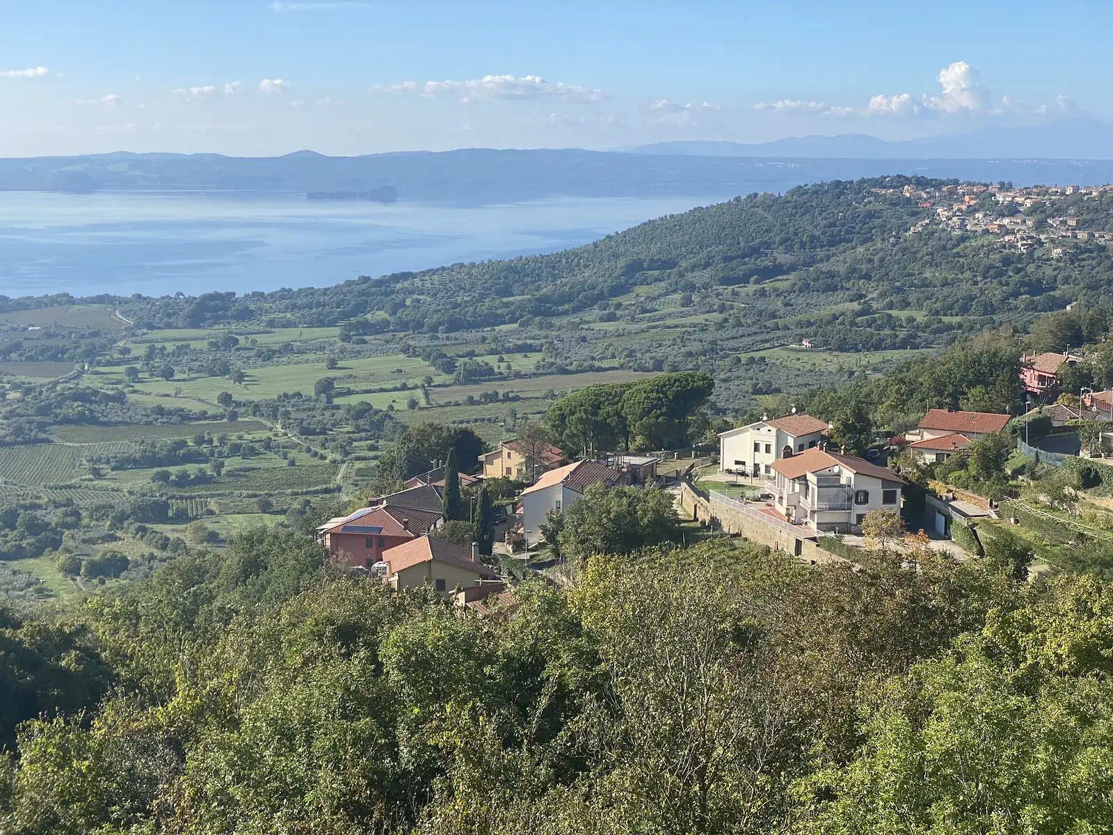 Terracotta-roofed houses sit on a wooded hillside overlooking a large lake and a wide green valley.