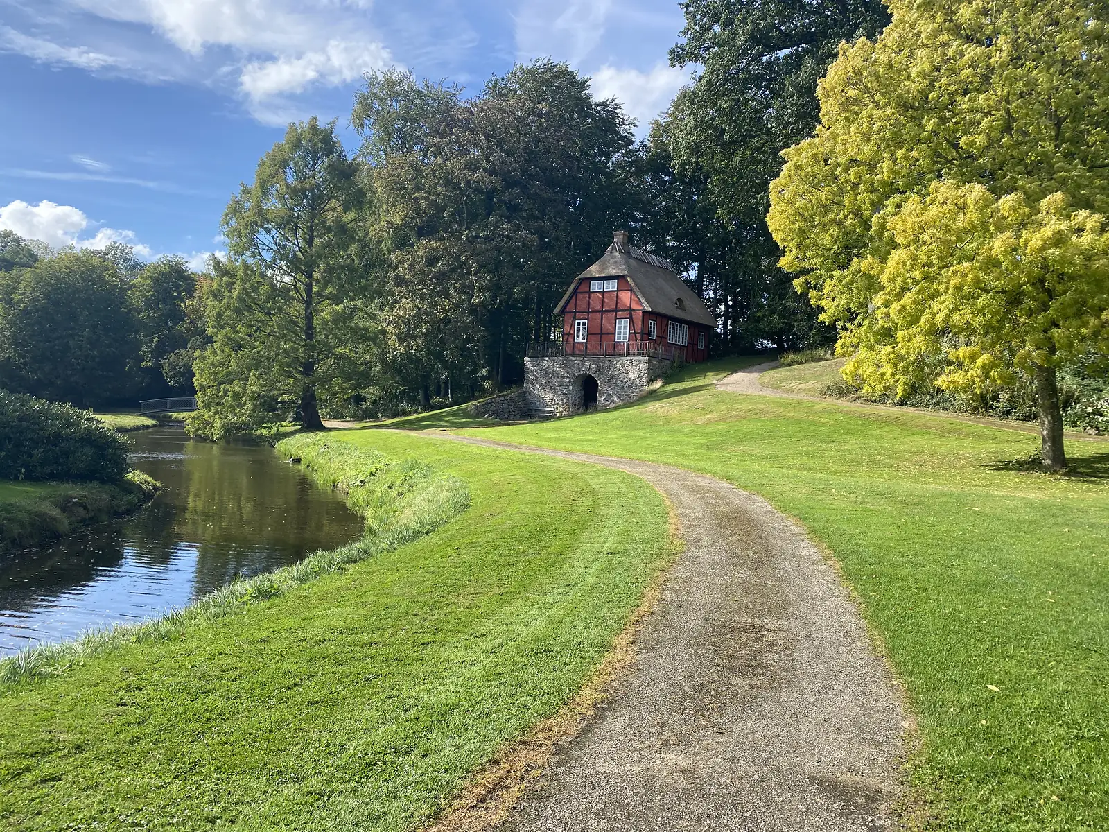 A red half-timbered thatched pavilion standing on a stone arched undercroft above a gravel path beside a canal.