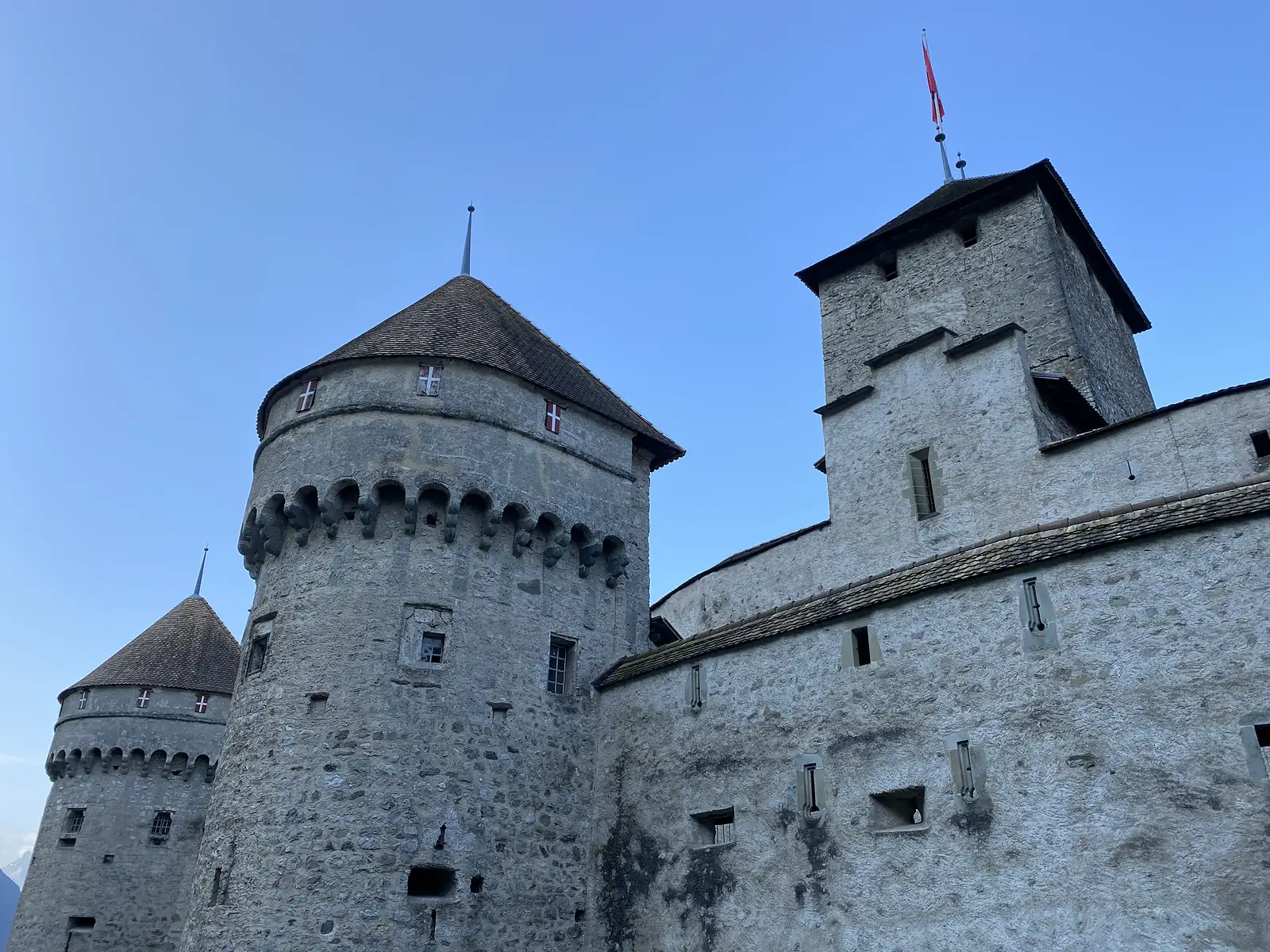 Round stone castle towers with conical slate roofs and small red-and-white shield motifs rise against a clear blue sky.