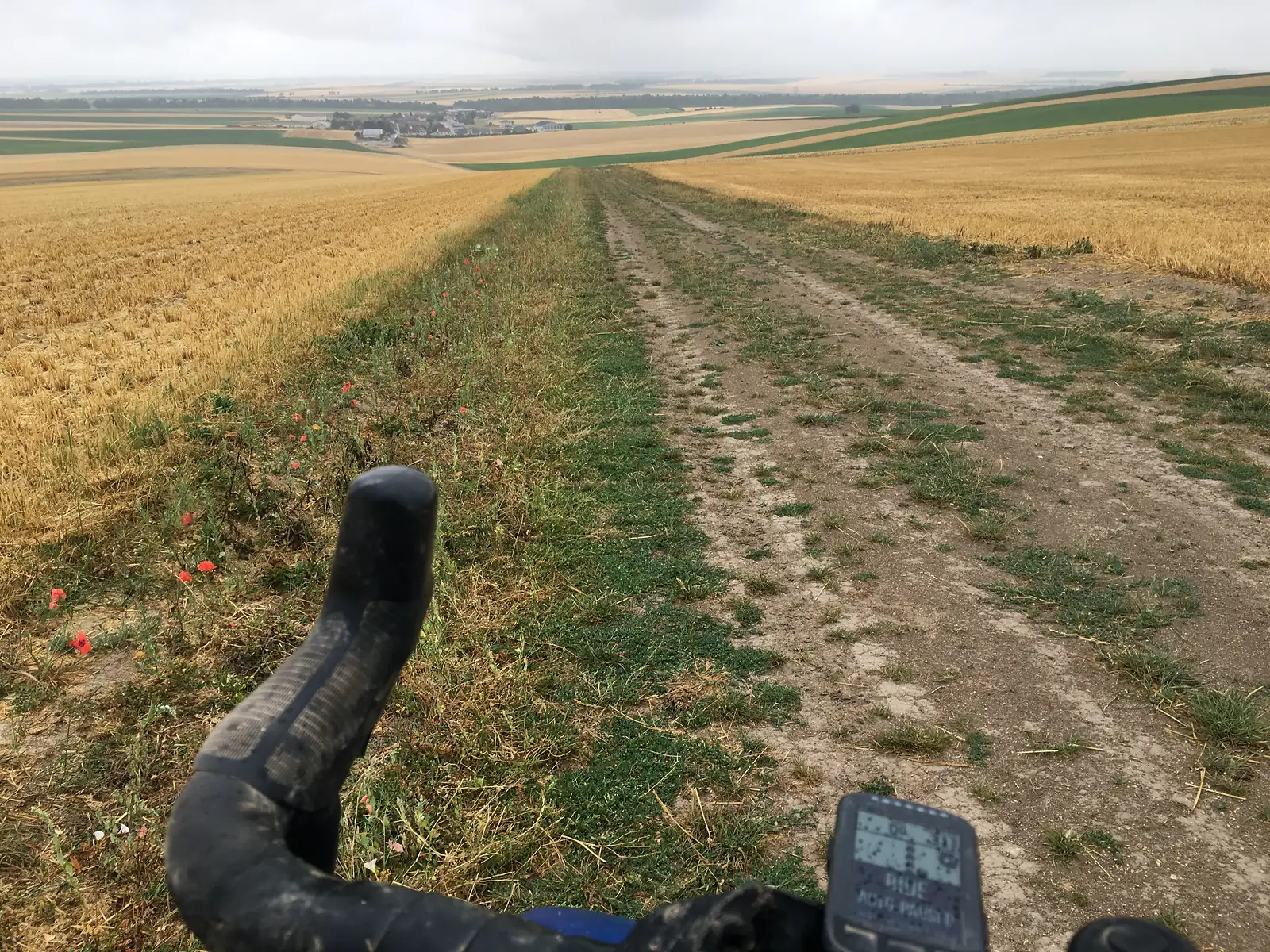 View over a bicycle's drop handlebars and cycling computer along a grassy farm track cutting between fields of harvested wheat, poppies growing at the field edge, with rolling countryside and a distant village beyond.