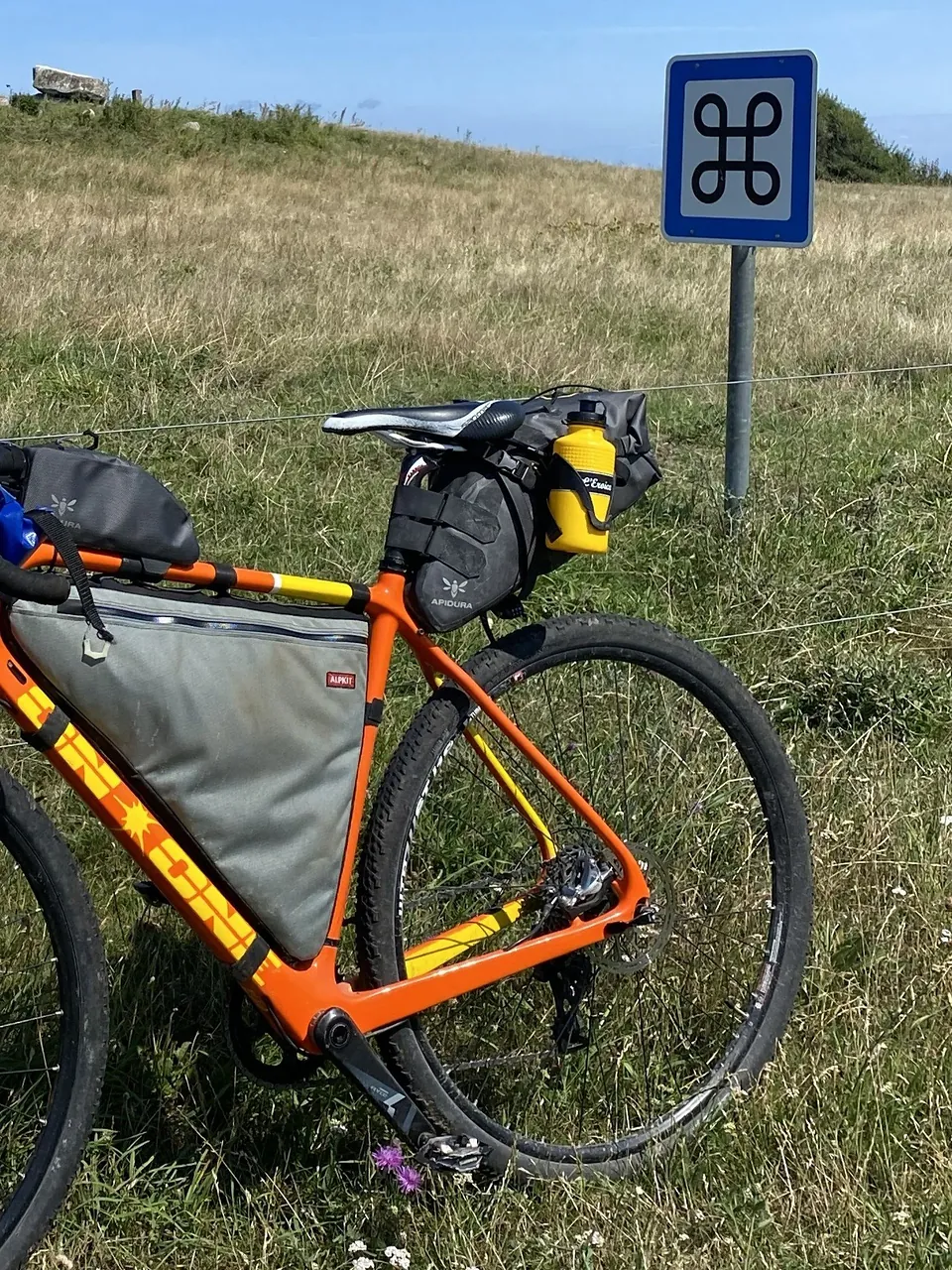 A orange bike with a grey frame pack, and a yellow water bottle on the side of the seat pack.