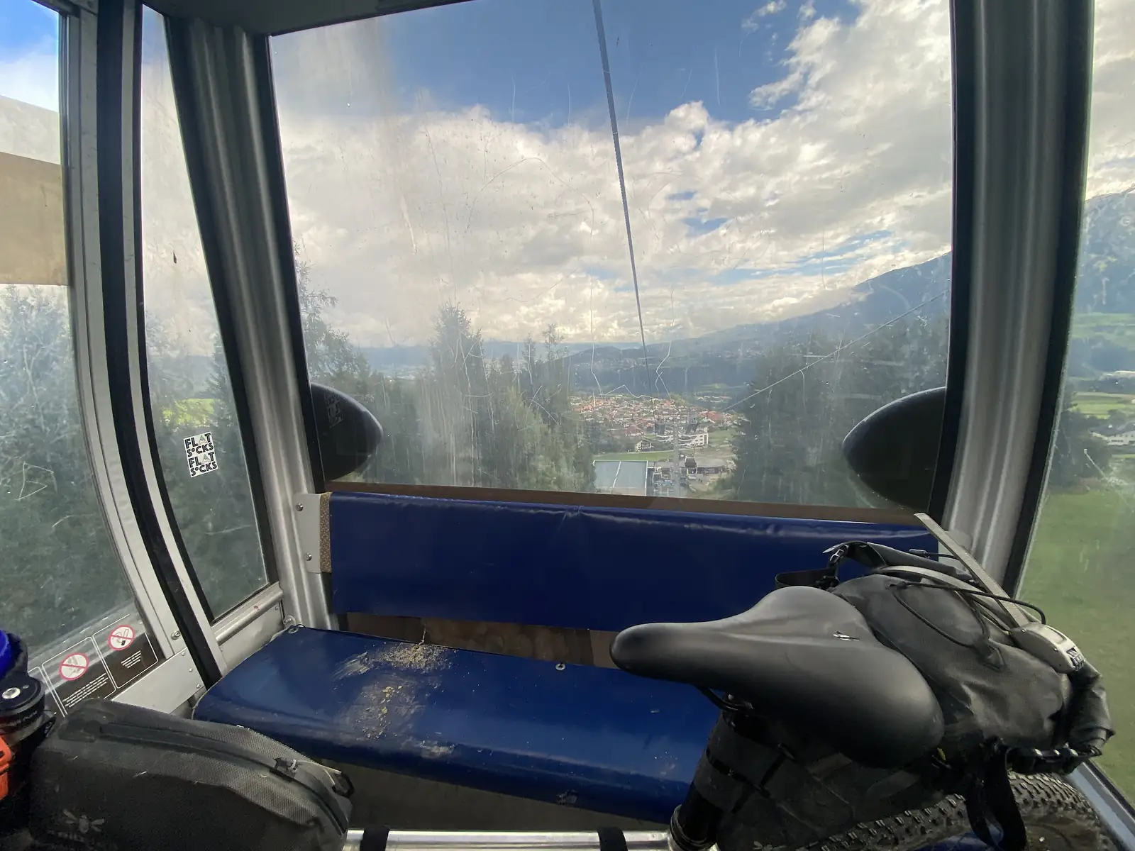 Inside a cable-car cabin with a blue bench seat, a loaded bike propped against it, looking down through scratched glass to a village in the valley far below.