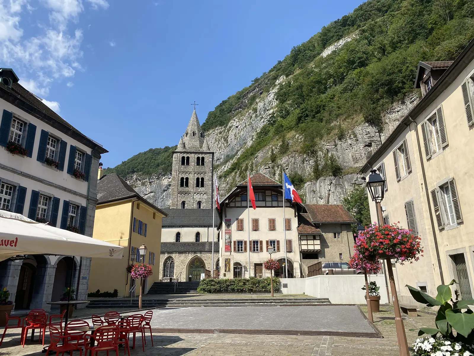 A Romanesque church bell tower rises above a paved square with café tables and hanging flower baskets, a sheer wooded cliff looming directly behind the buildings.