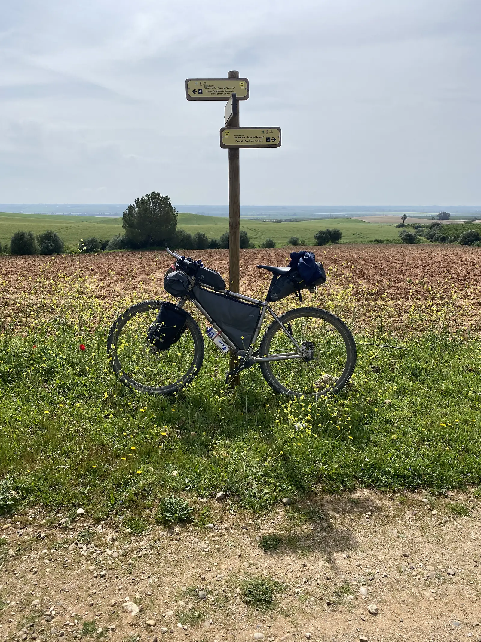 A loaded bikepacking bike propped against a waymark post signed "Sierrezuela - Rozas del Pozuelo", on a dirt track between a ploughed field and green cereal.