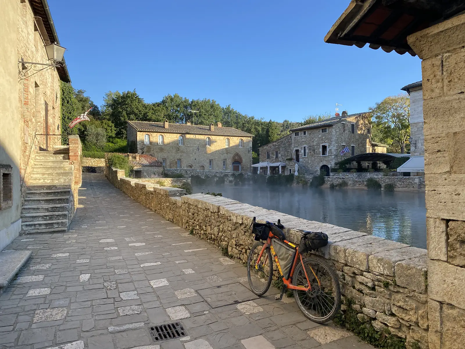 A loaded orange bikepacking bike resting against a low stone wall beside a steaming thermal pool, with stone village buildings around the water.