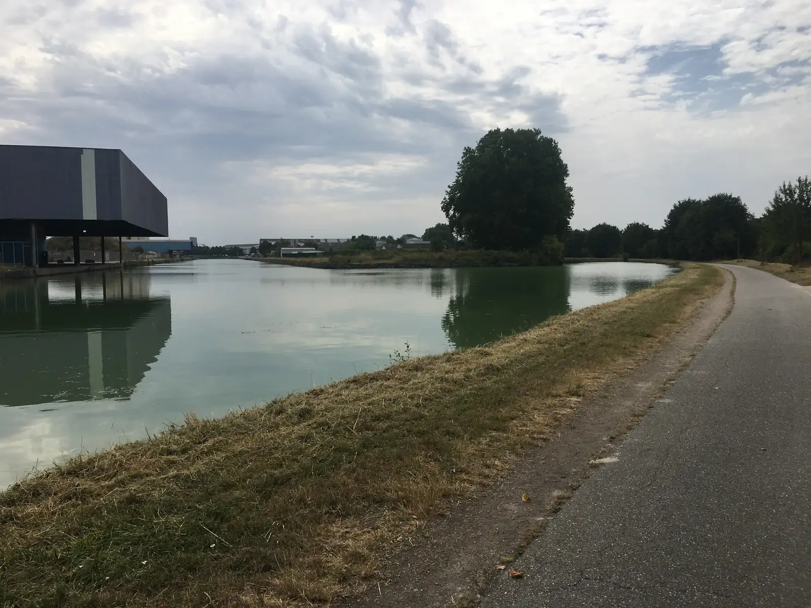 A canal reflecting an overcast sky beside a paved towpath, an industrial shed on one bank and a large tree marking a bend on the other.