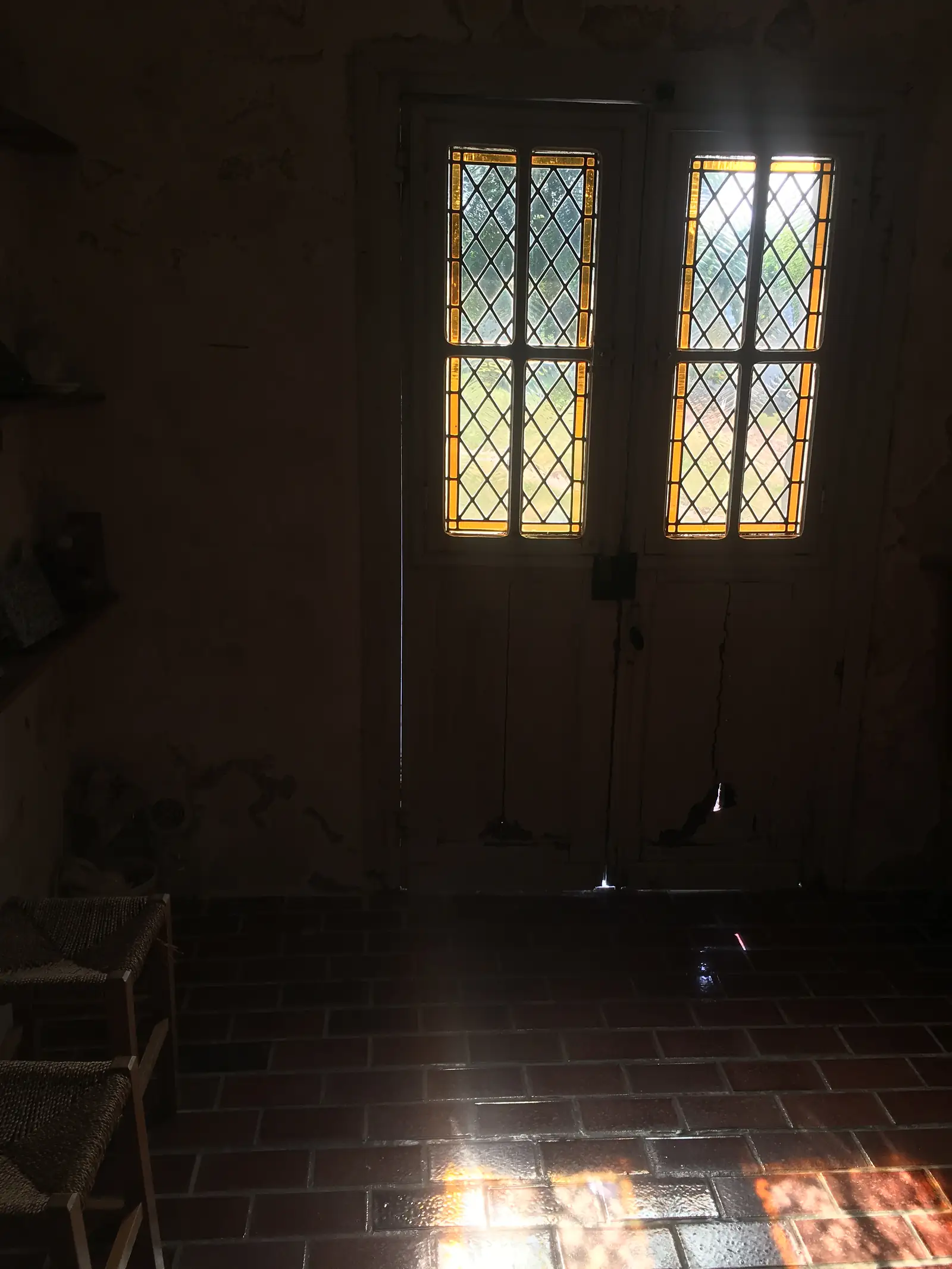 Sunlight glows through two small diamond-leaded windows in the dim, timber-lined interior of an old building, its walls patched and peeling.