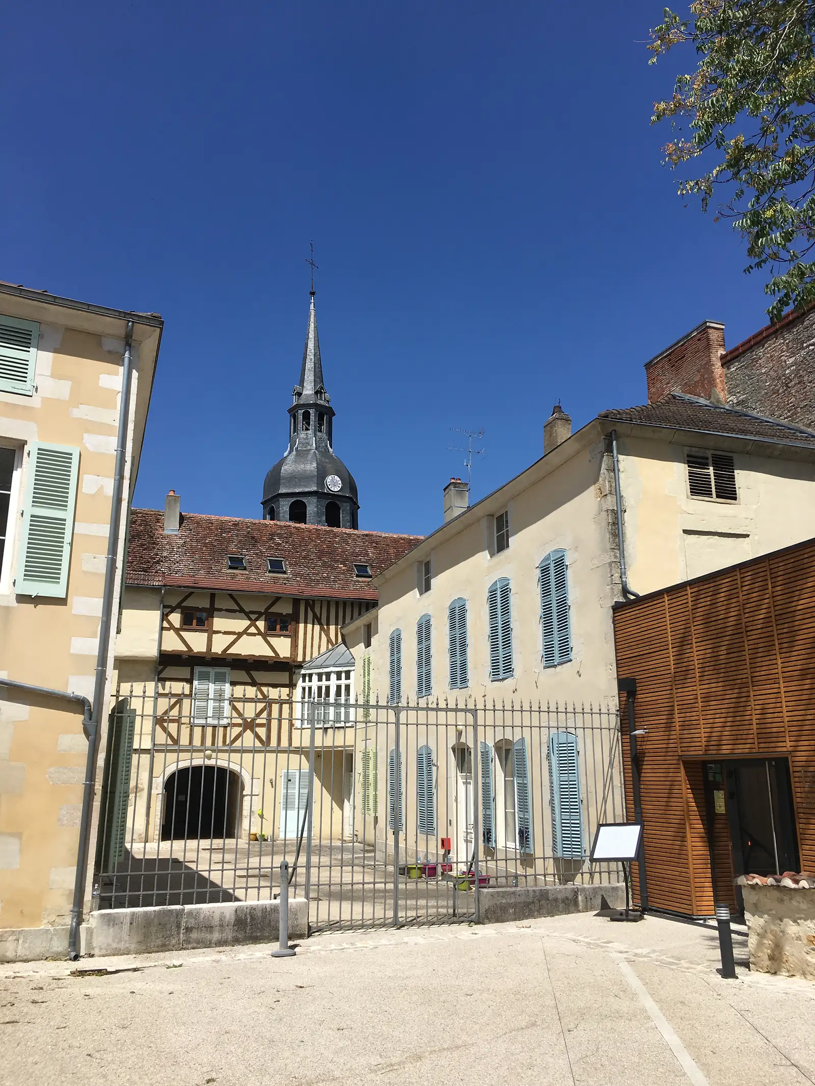 A slate-covered clock tower and spire rise above a half-timbered building on a village square, seen against a cloudless sky.