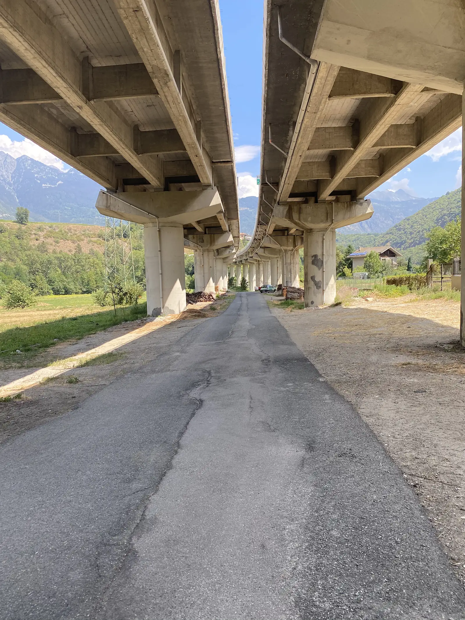 A gravel lane runs beneath twin elevated motorway viaducts supported on tall concrete piers, mountains visible through the gap between the two carriageways.