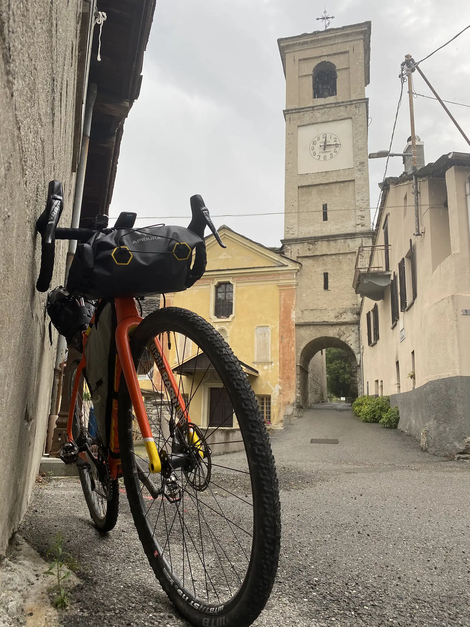 A loaded orange gravel bike stands in a narrow alley beneath a stone clock tower marked 'COMUNE STRAMBINO', with a yellow church and archway behind.