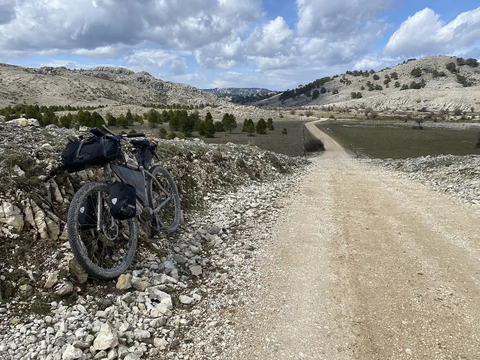 A loaded bikepacking bike propped on a bank of white limestone rubble beside a pale track winding down into a juniper-dotted basin.