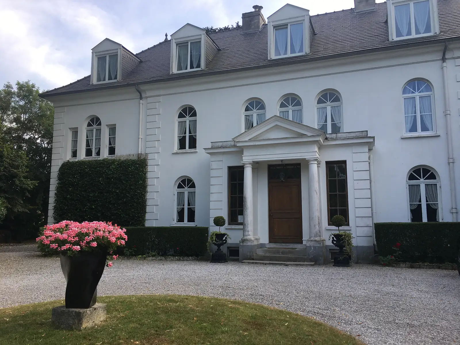 The white stuccoed front of a grand manor house with an arched-window facade and a columned entrance porch, a pot of pink flowers on the gravel drive.