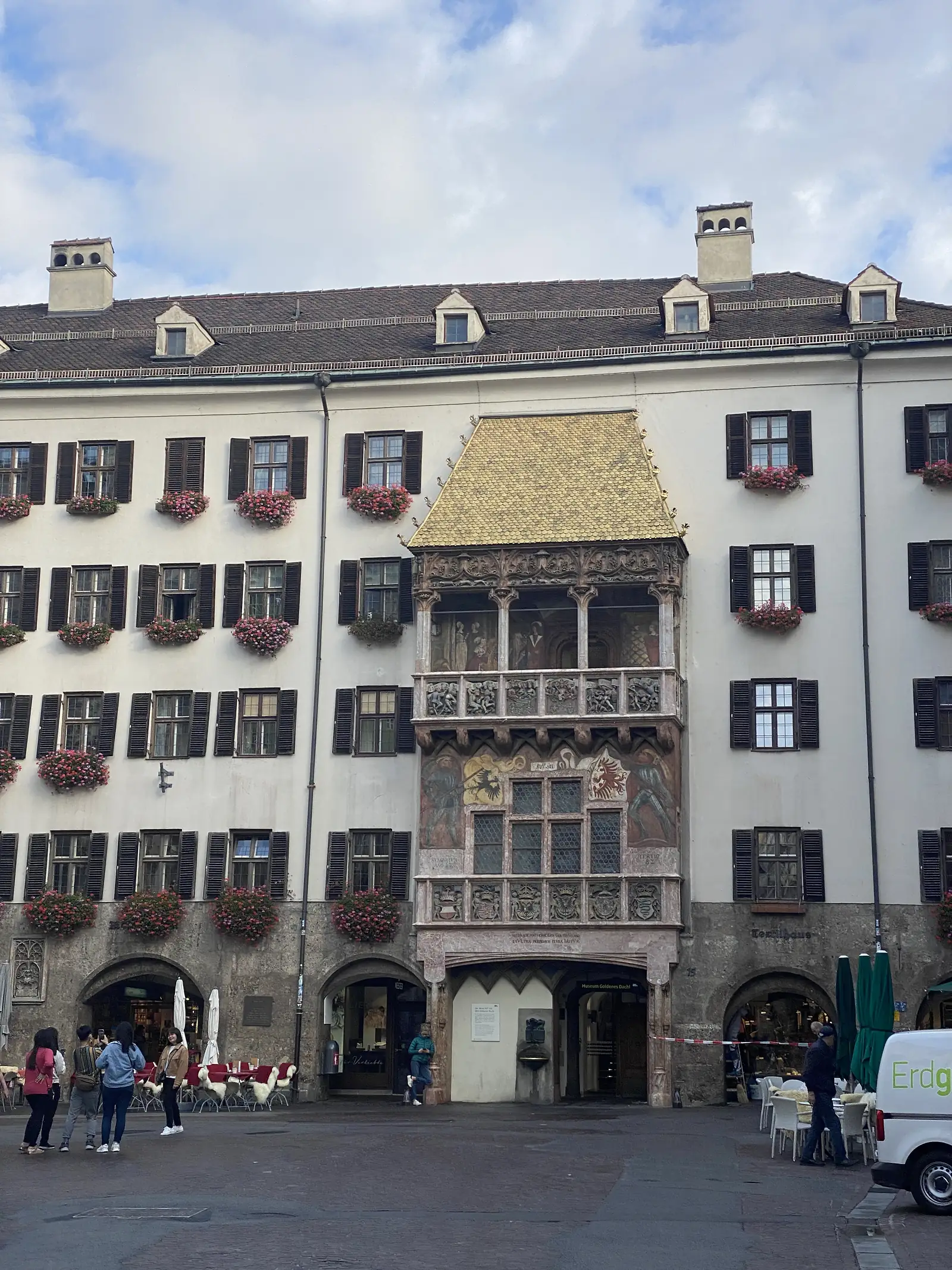 A grand townhouse facade with dark-shuttered windows overflowing with pink flowers, dominated by a central gabled bay with a gilded, scale-tiled roof above a richly painted and carved oriel featuring coats of arms, with cafe tables and umbrellas set out on the square below.