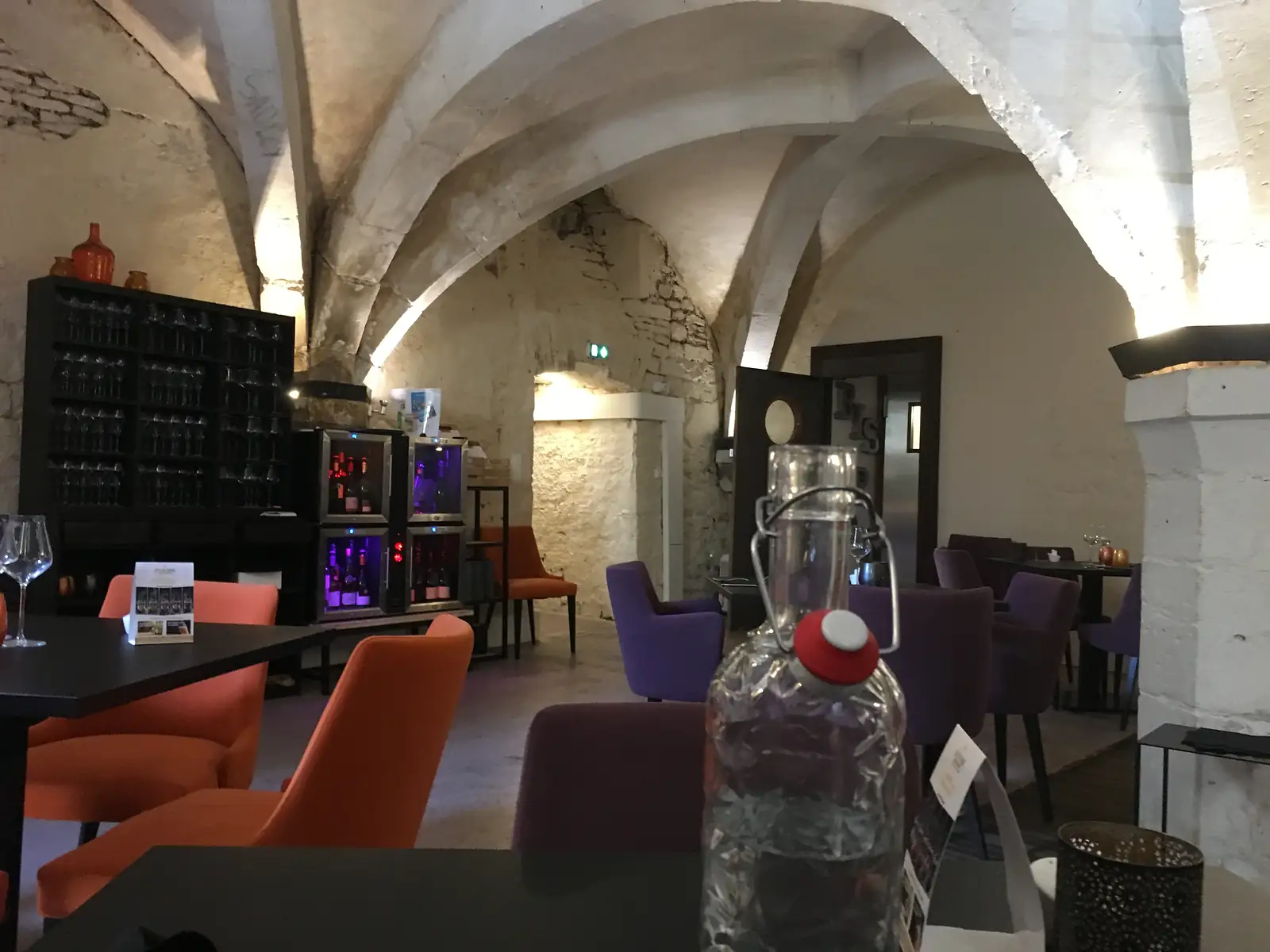 A vaulted stone-ceilinged cellar bar furnished with orange and purple armchairs, wine glasses and a large glass water carafe on the table.