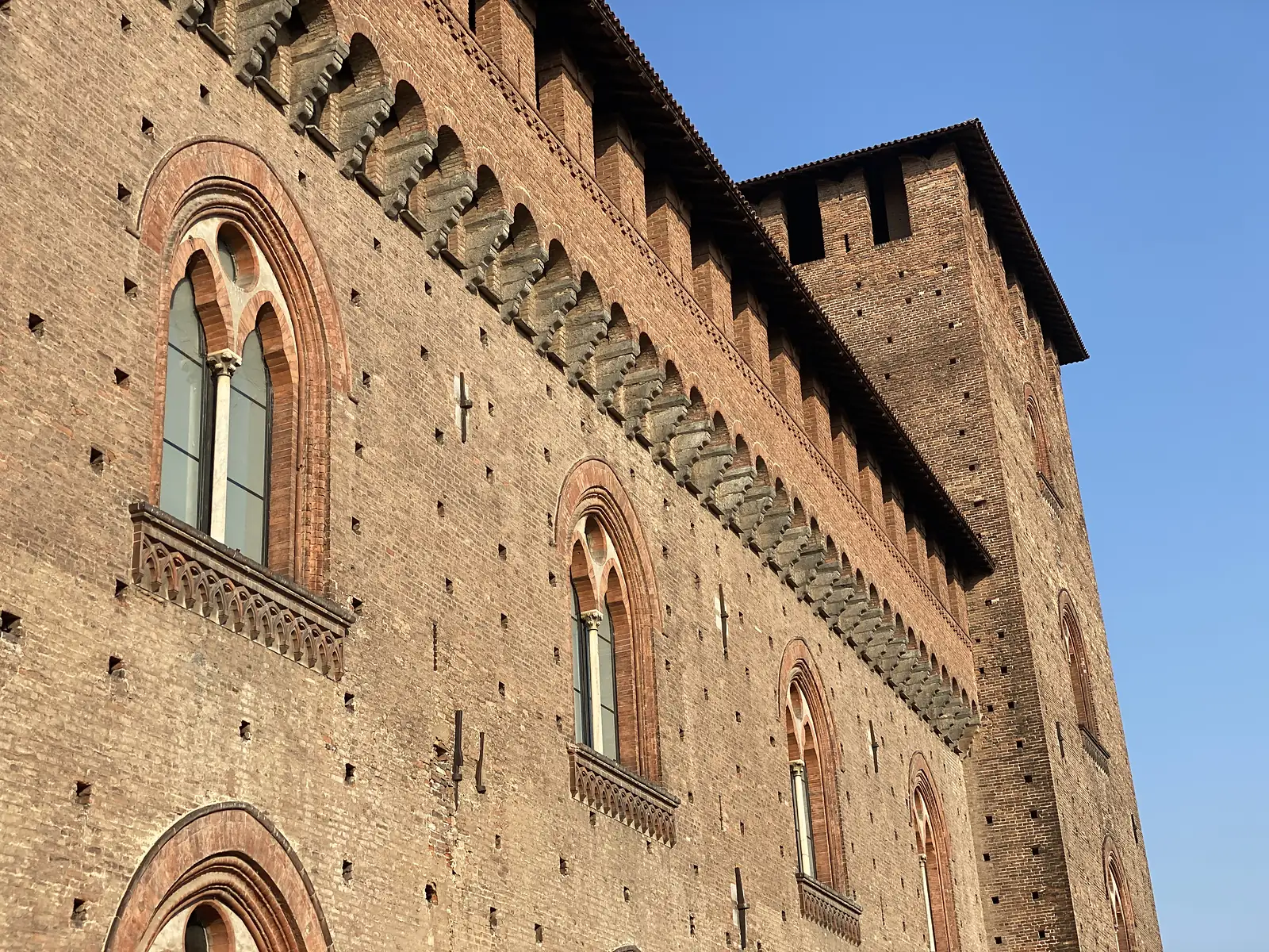 Gothic arched windows with slender colonnettes line the brick façade of a fortified palace beneath a corbelled, crenellated parapet.
